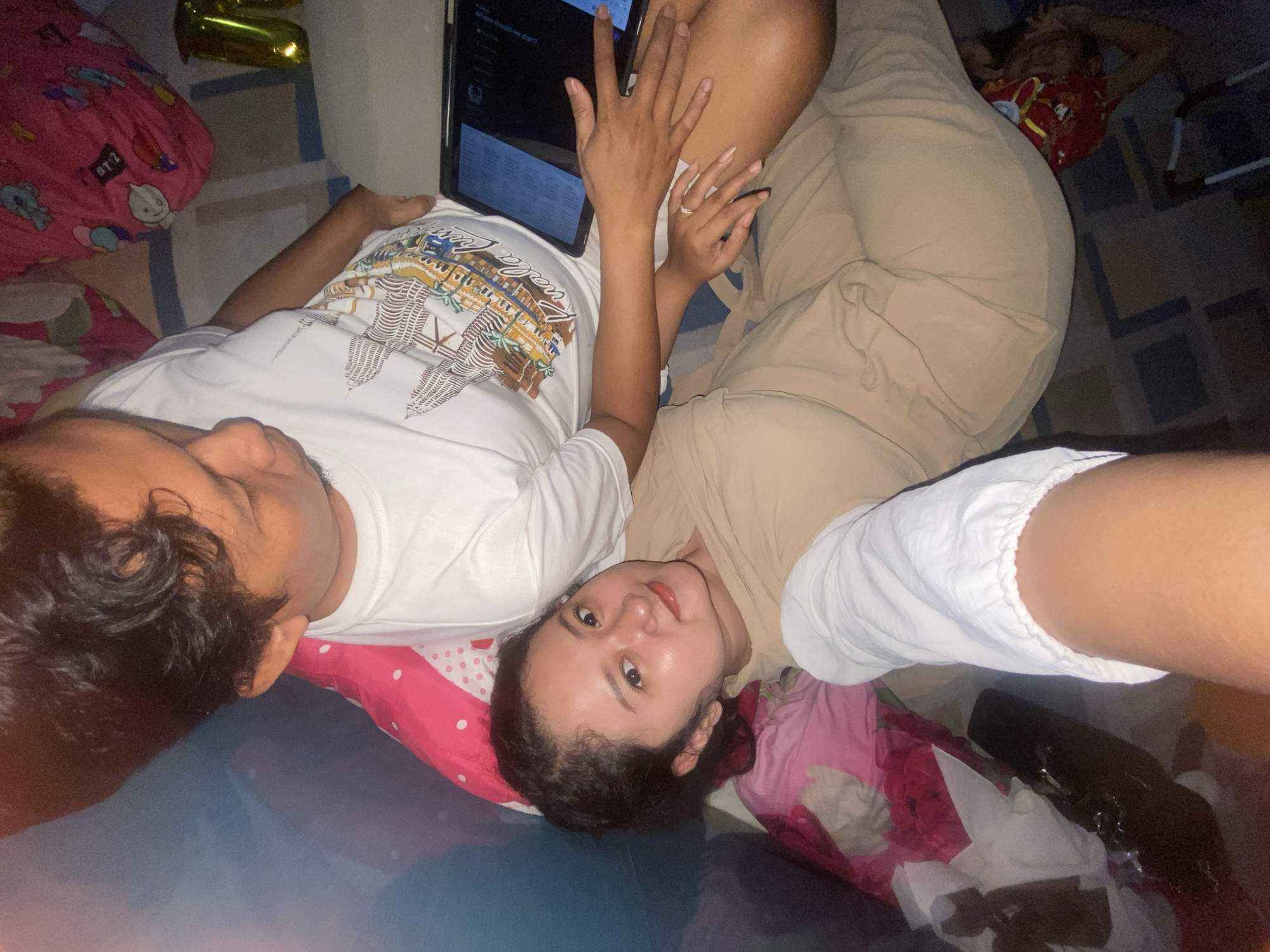 Two individuals are positioned on the floor of a room. One person is reclining on a red-and-white patterned pillow, while the other is lying adjacent with a tablet positioned on their midsection. The person reclining appears to be looking at the camera, while the tablet screen displays an open application. There is another person reclining further back in the image. The background consists of a tiled floor and assorted objects, including a mylar balloon. The location is presumed to be in Gelam, Indonesia.