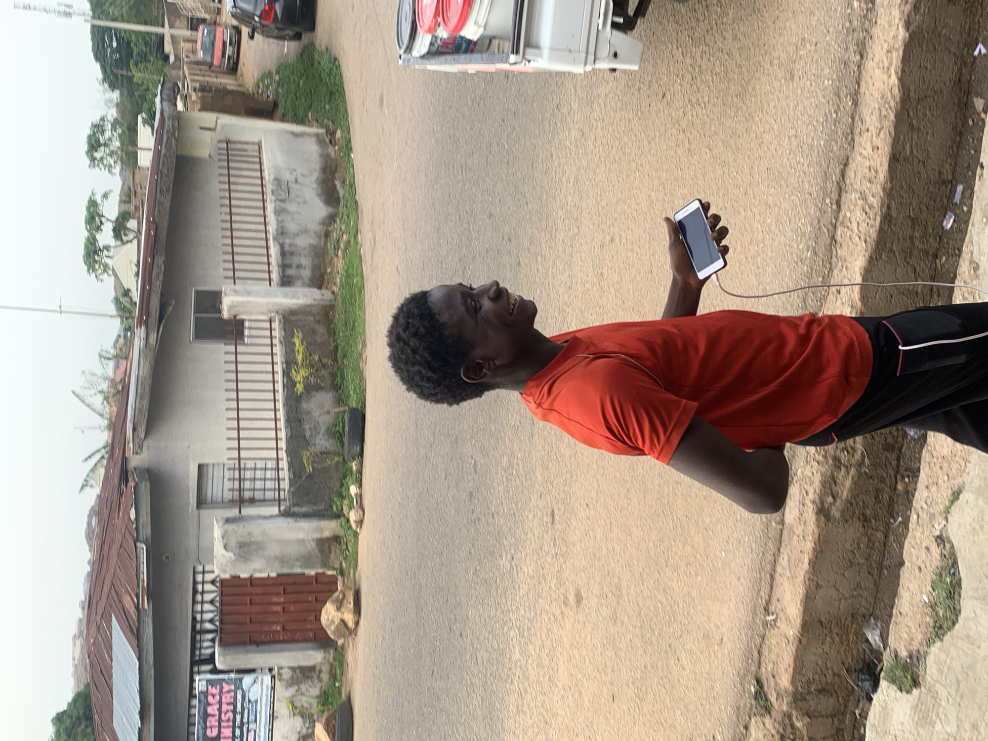 A young Black man is standing on a paved road in Barakin Akawo, Nigeria. He is wearing a red t-shirt and black pants with a white stripe down the side. He is holding a smartphone in his right hand, which is connected to a charging cable. The road has a sidewalk on one side, with a building and foliage visible in the background. A vehicle is partially visible at the top of the frame. The image is slightly rotated. A sign saying "GRACE MISTRY" can be seen on the side of the building.
