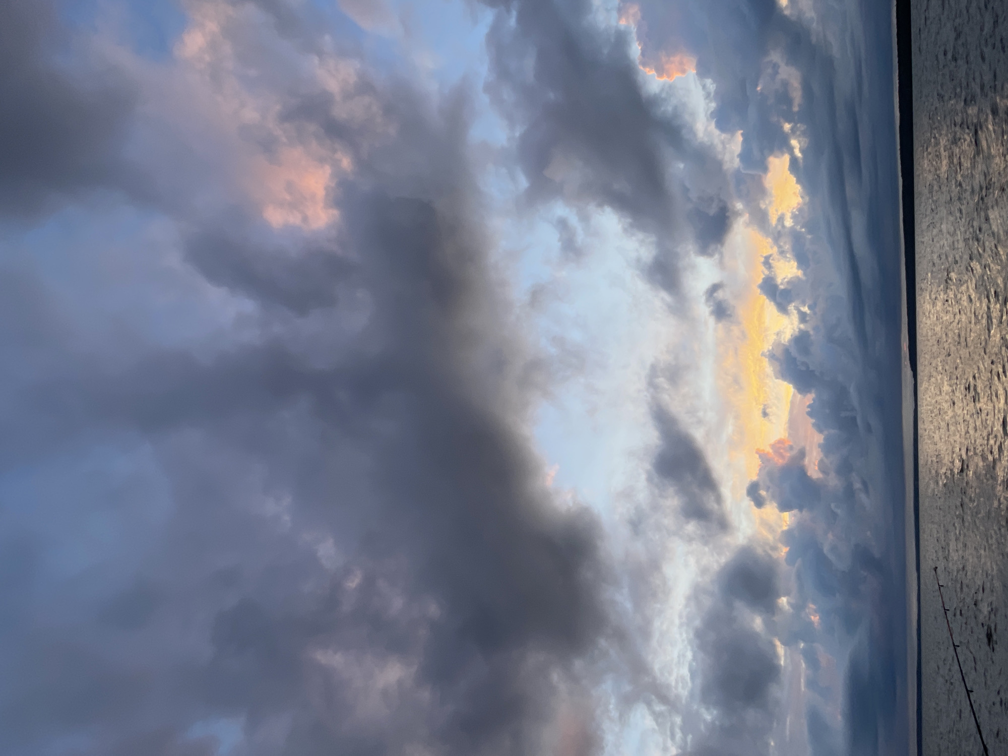 An eye-level, full shot captures a body of water under a dramatic, cloudy sky at dusk. The water, appearing dark and choppy, reflects the muted colors of the sky, with hints of orange and pink breaking through the dominant grays and blues. A dark, silhouetted landmass is visible on the horizon. In the foreground, a fishing rod rests on the water, its tip extending slightly into the frame. The sky is filled with large, textured clouds, some tinged with the warm glow of sunset. The scene evokes a sense of calm and solitude. The image is taken in Yablonovka, Russia.