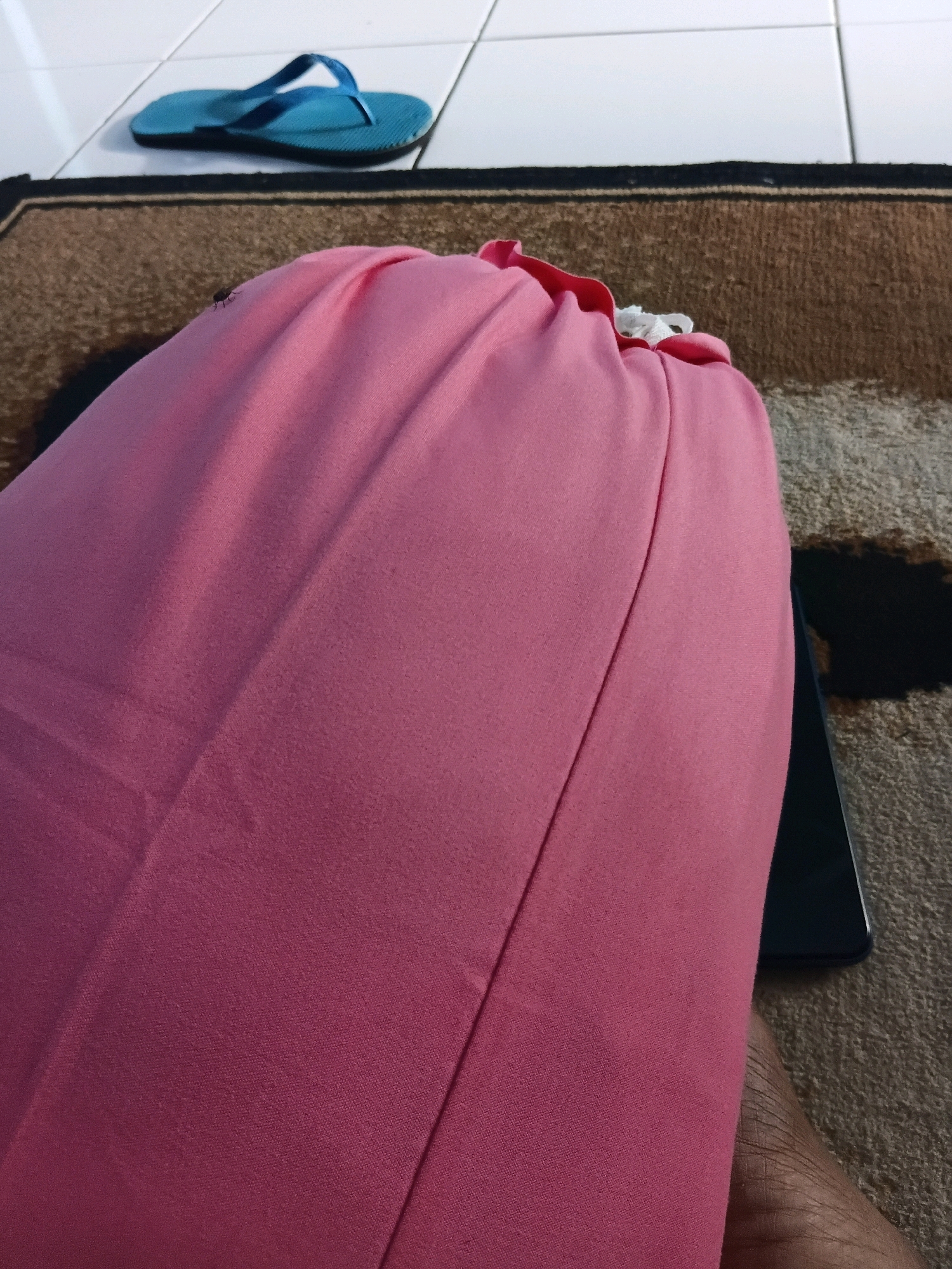 In the city of Tanggul Wetan, Indonesia, a close-up shot captures a person's leg, covered by a pink garment, resting on a carpeted floor. To the left, a blue flip-flop rests on a tiled surface. A dark, rectangular object, possibly a phone, is partially visible beneath the pink fabric. A small fly is perched on the pink garment, adding a detail of insect life within the domestic scene. The lighting suggests an indoor environment, possibly a home, with the focus on the textured surfaces of the carpet and fabric.
