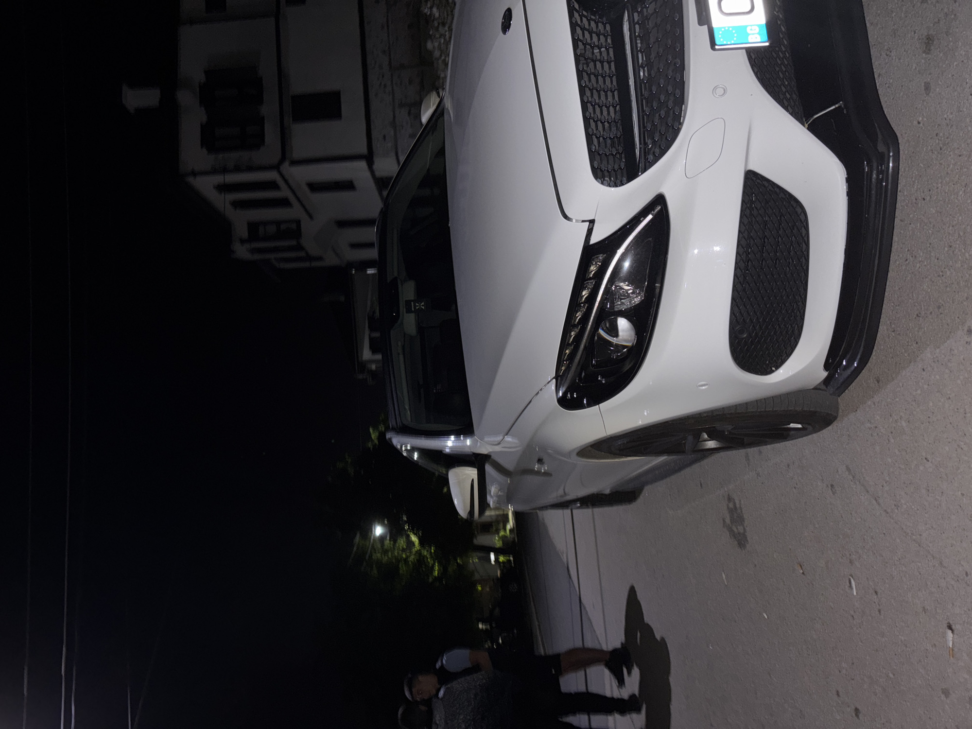The image captures a nighttime street scene in Zlatograd, Bulgaria. A white Mercedes-Benz sedan, appearing to be a C-Class model, is prominently positioned on the right side of a paved road. The vehicle features modern LED headlights, a black grille, black multi-spoke wheels on the front-right, and an aftermarket black front splitter attached to the lower bumper. A Bulgarian license plate, displaying the "BG" country code, is partially visible on the front bumper. An air freshener is seen hanging from the interior rearview mirror.
Two male individuals are situated on the left side of the street. The leftmost person is viewed from the back, wearing a dark, possibly patterned, top and dark trousers. The second individual, to the right of the first, wears a light-colored top and dark shorts, and appears to be facing the first person. Their shadows are cast towards the left side of the frame on the asphalt surface, which shows scattered small debris, possibly cigarette butts.
In the mid-ground, a street light emits a bright illumination, revealing the dark foliage of trees. The background features a multi-story, white architectural structure with dark window frames and a chimney, indicative of traditional Bulgarian building styles. The sky is dark, with thin overhead power lines visible at the very top of the image.