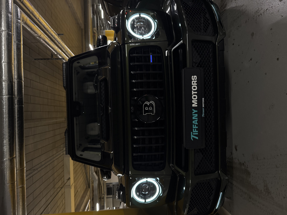 A black Brabus SUV dominates the frame, parked inside what appears to be a parking garage. The vehicle's front grille features the signature "B" emblem. The "Tiffany Motors" branding is visible on the car. One door is open, revealing the interior. The background is a dimly lit parking structure with pipes and concrete walls. The image is rotated.