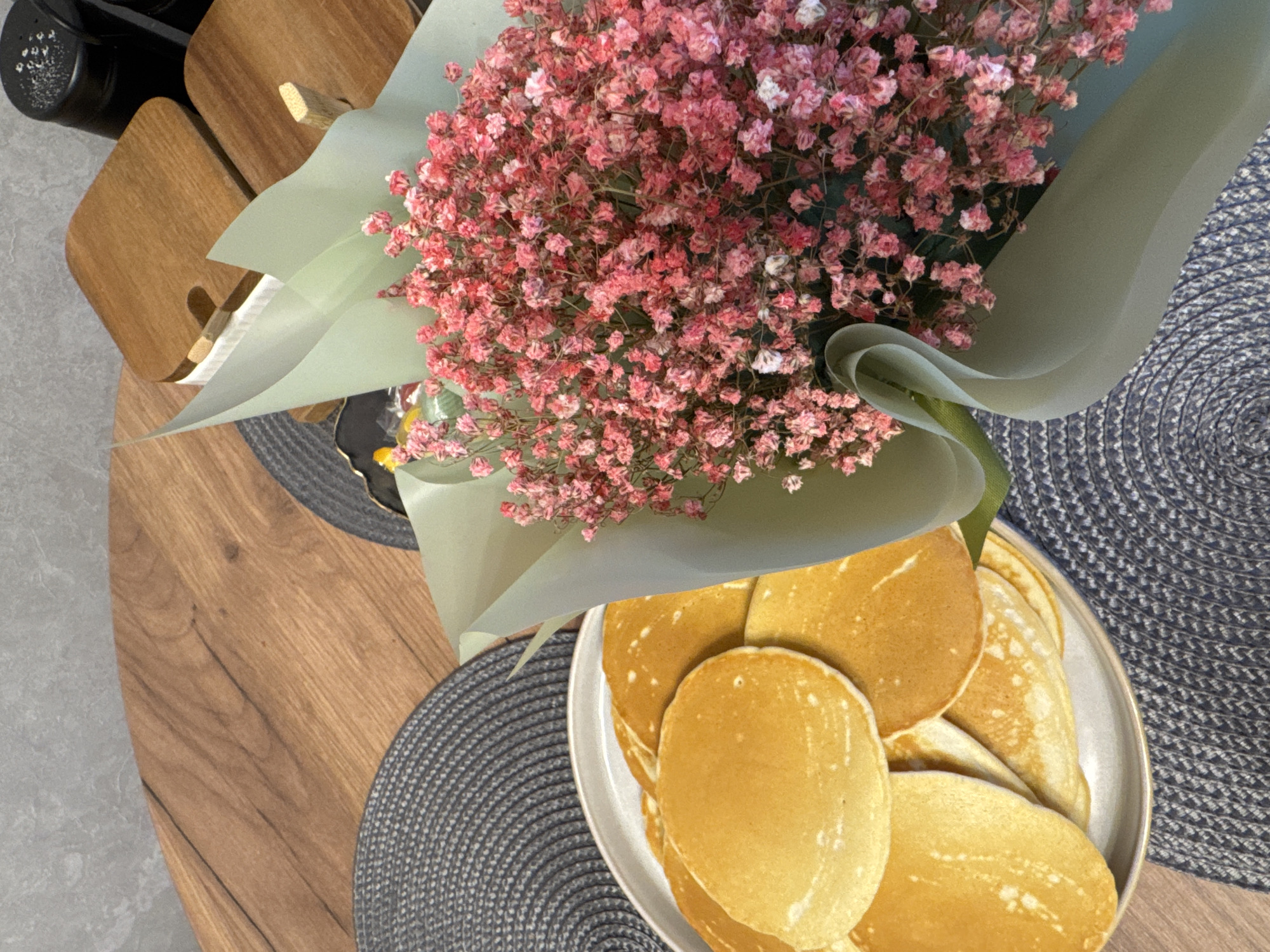 A high-angle shot displays a scene on a wooden table in Yakhroma, Russia. A bouquet of pink baby's breath flowers, wrapped in light green paper, dominates the center of the frame. To the right of the bouquet, a white plate holds a stack of golden-brown pancakes. Underneath both the bouquet and the plate of pancakes are dark gray, round placemats with a woven texture. To the left of the bouquet, wooden objects, possibly coasters or small cutting boards, are stacked next to a dark-colored object, possibly a container. The table surface has a wood grain pattern.