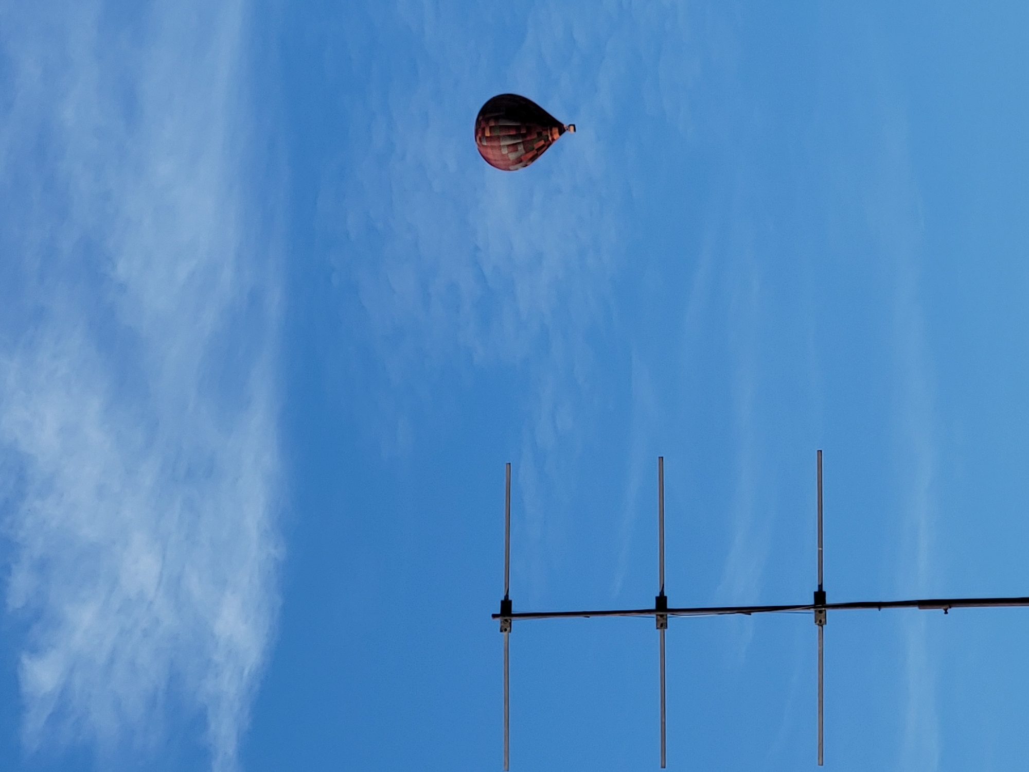 An outdoor, eye-level shot shows a deep blue sky with wispy white clouds. A hot air balloon with red and gray panels is visible in the upper right quadrant of the frame. In the lower left quadrant, a tall antenna with three horizontal crossbars extends upwards. The scene is devoid of any people or specific landmarks, making its location in "Unknown, Unknown" plausible.