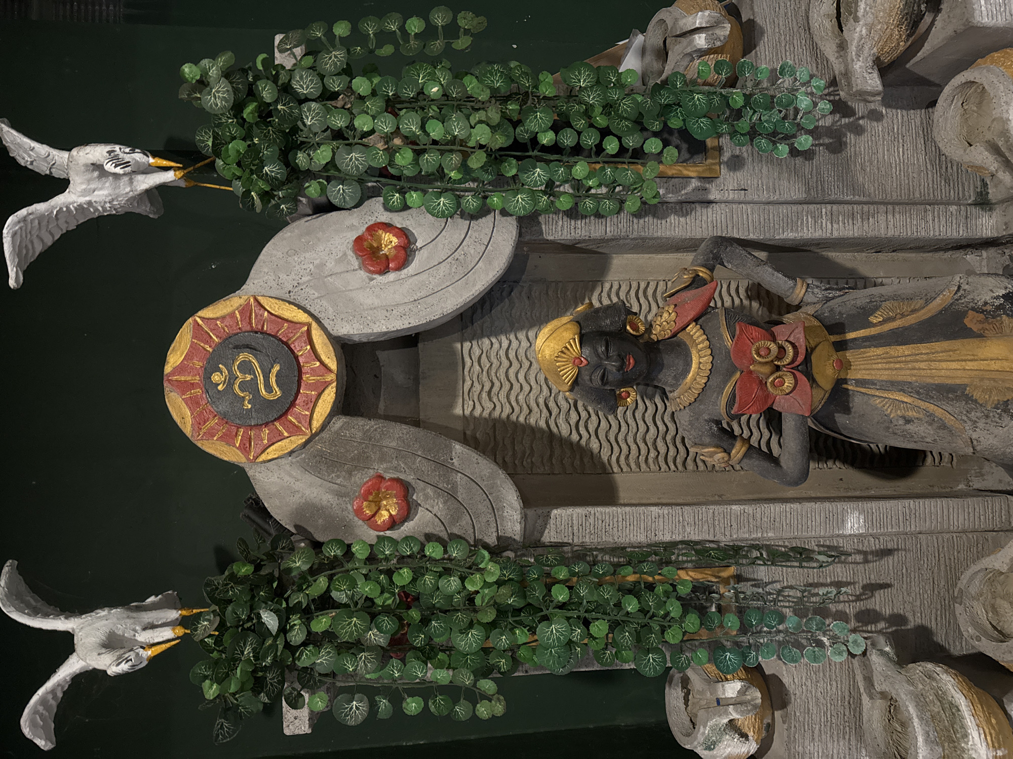 The image depicts an ornate statue of a Balinese woman, likely a dancer, positioned centrally within a stone structure. She is adorned with traditional clothing and jewelry, including a golden headdress, a red and gold flower ornament on her chest, and a necklace. Her skin is dark, and she wears red lipstick.
Flanking the central figure are two grey, stylized bird statues, with wings outstretched. They appear to be perched on top of decorative stone elements. Cascading artificial green vines with small, circular leaves hang down from these elements on either side of the central statue, adding a touch of greenery.
Behind the statue, a large, circular ornament is visible, framed by stone arches. This ornament features a red and gold border and a dark center with a golden symbol that resembles the "Om" mantra from Hindu and Buddhist traditions. The stone structure itself is detailed with carved patterns, including wavy lines behind the central statue.
The overall aesthetic suggests a cultural or artistic display, possibly found in a temple, garden, or decorative installation. Given the context of Kuta, Indonesia, this artwork likely represents Balinese culture and spirituality. The lighting is somewhat dim, creating shadows that highlight the textures of the stone and the statues. The background is a dark, solid color, likely a wall.