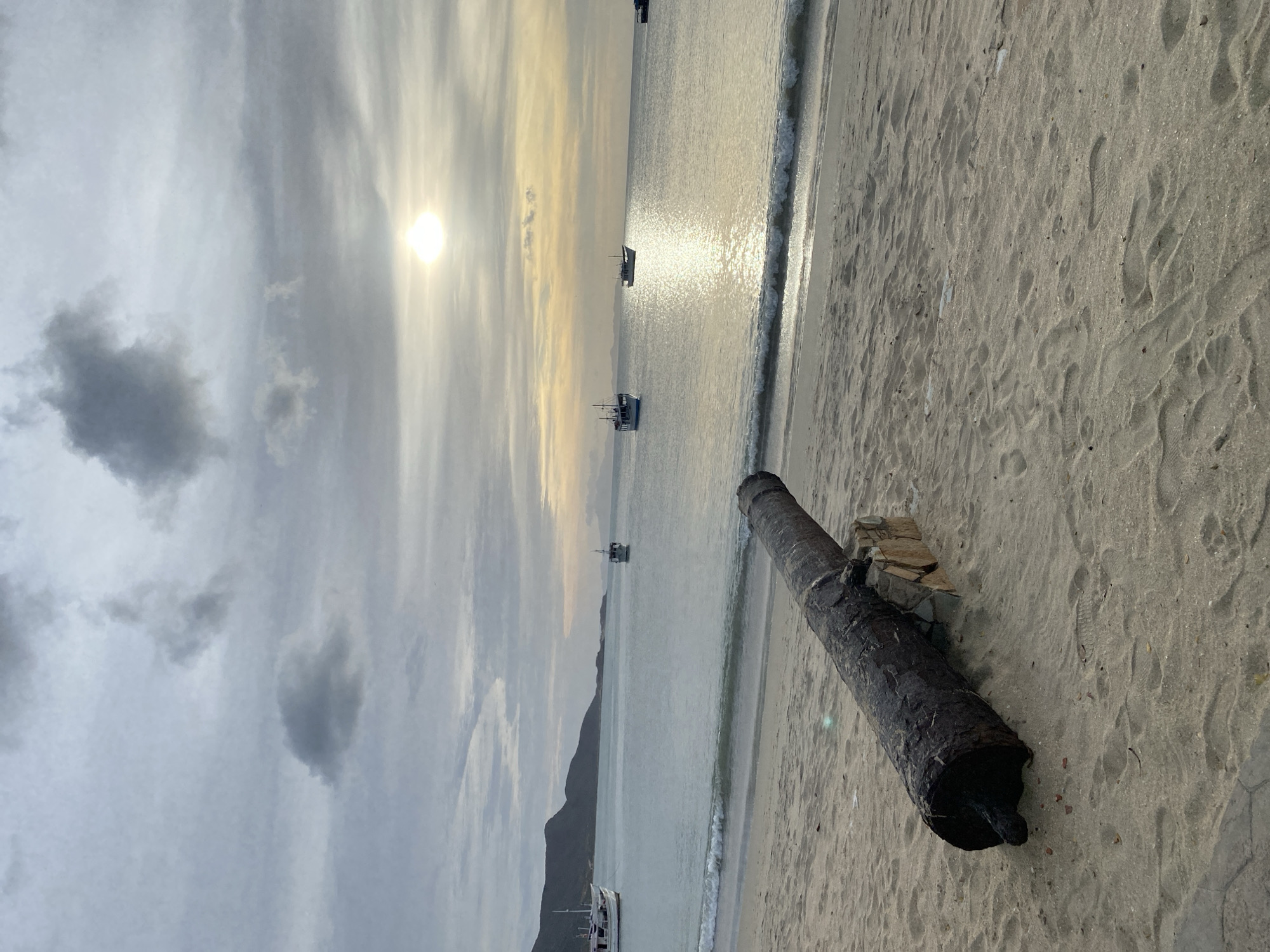 The scene depicts a coastal area in Juan Griego, Venezuela, under an overcast sky with the sun positioned low on the horizon, creating a bright reflection path across the water. In the foreground, a dark, weathered metal cannon barrel rests horizontally on light brown sand, supported by a structure of unmortared, rough-cut stones. The sandy beach contains multiple footprints and minor ripples near the waterline. Small waves break gently onto the shore. The ocean extends into the background, occupied by at least five small to medium-sized boats, one of which is a white vessel with blue trim on the far left. A range of dark hills or mountains forms the landmass along the left side of the background horizon. No individuals are present within the frame.