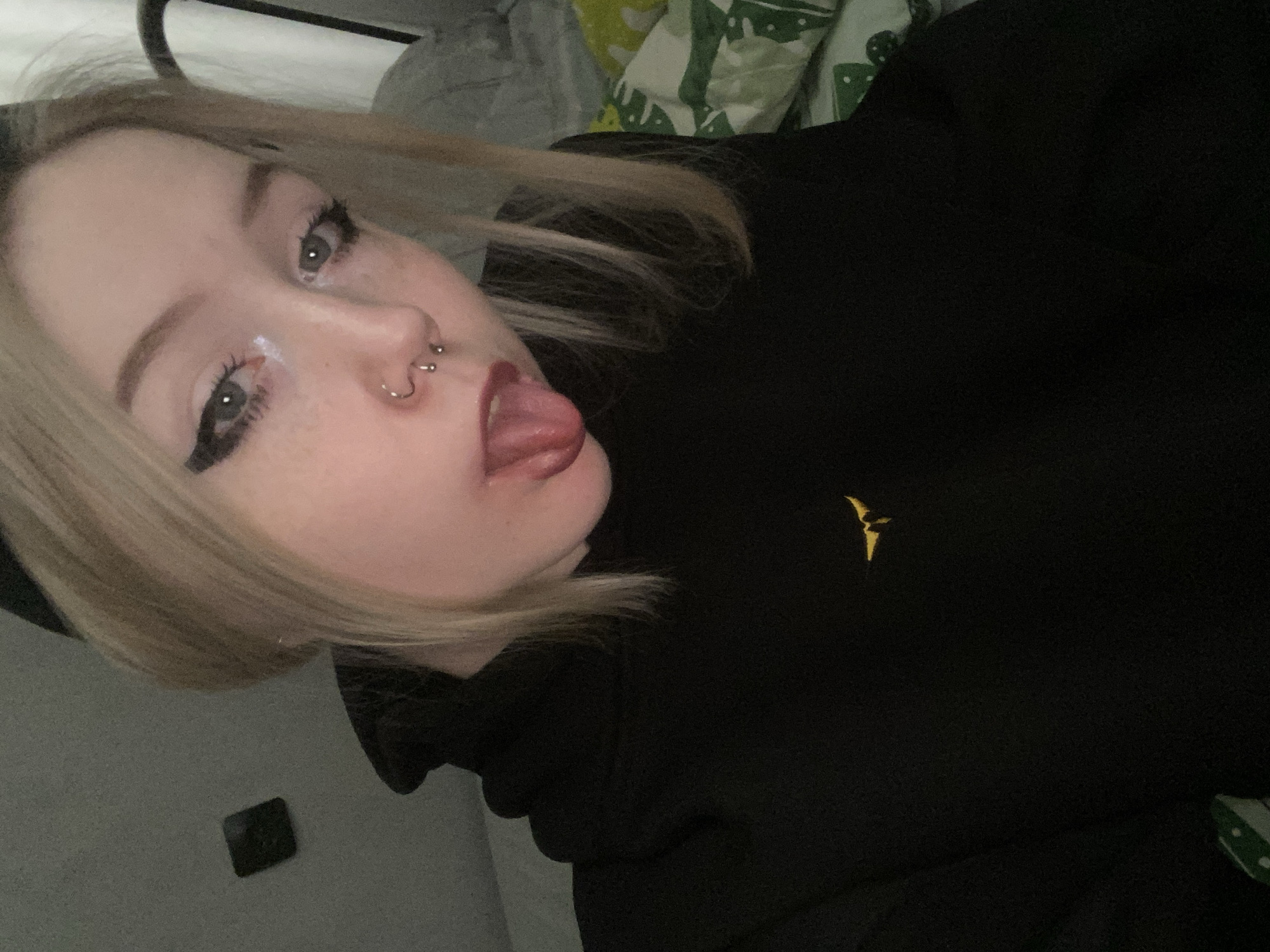 A selfie taken by a fair-skinned young woman with blonde hair, gray eyes, and visible makeup including eyeliner and a glossy substance on her face. She has piercings on her nose and is sticking out her tongue. She is wearing a black hoodie with a yellow bird emblem on the right chest. The background appears to be a room with a bed. There's a gray-colored object and a green and white patterned object partially visible in the background. The lighting is dim. The city of Izhevsk, Russia is not discernible in the image.