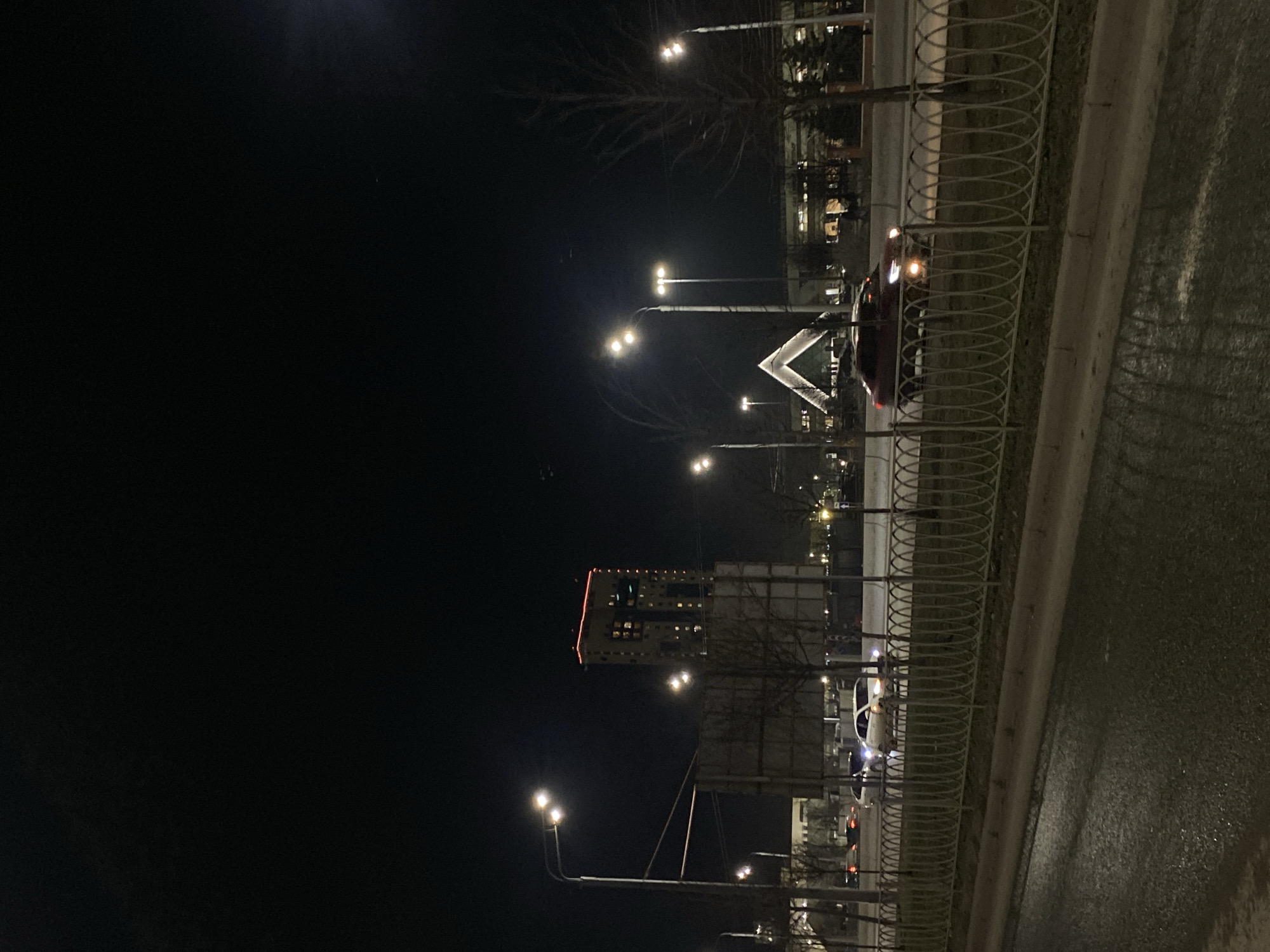 The image shows a night scene in Kazan, Russia. It features a road with a metal fence on one side, partially obscuring a row of street lights and buildings in the background. A car with its headlights on is visible behind the fence. The building has a distinct shape, possibly a modern architectural design. Other buildings and street lights are seen further in the background. The sky is dark, indicating it is night.