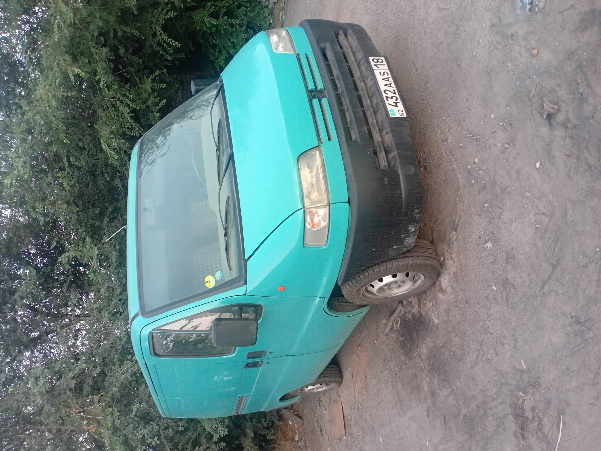 An abandoned turquoise van, identified by its license plate "KZ 432 AAS 18," is parked on a dirt road in a wooded area on the outskirts of Semey, Kazakhstan. The van, appearing to be a panel van, is the sole subject of the image, positioned at an angle facing the viewer. The foreground consists of a dusty, uneven dirt surface with scattered debris, including small stones and pieces of wood. In the background, dense green foliage from trees and bushes forms a natural backdrop. The van's condition suggests it has been stationary for some time, with dust and dirt covering its surfaces. The lighting indicates it is either early morning or late afternoon, with soft shadows.