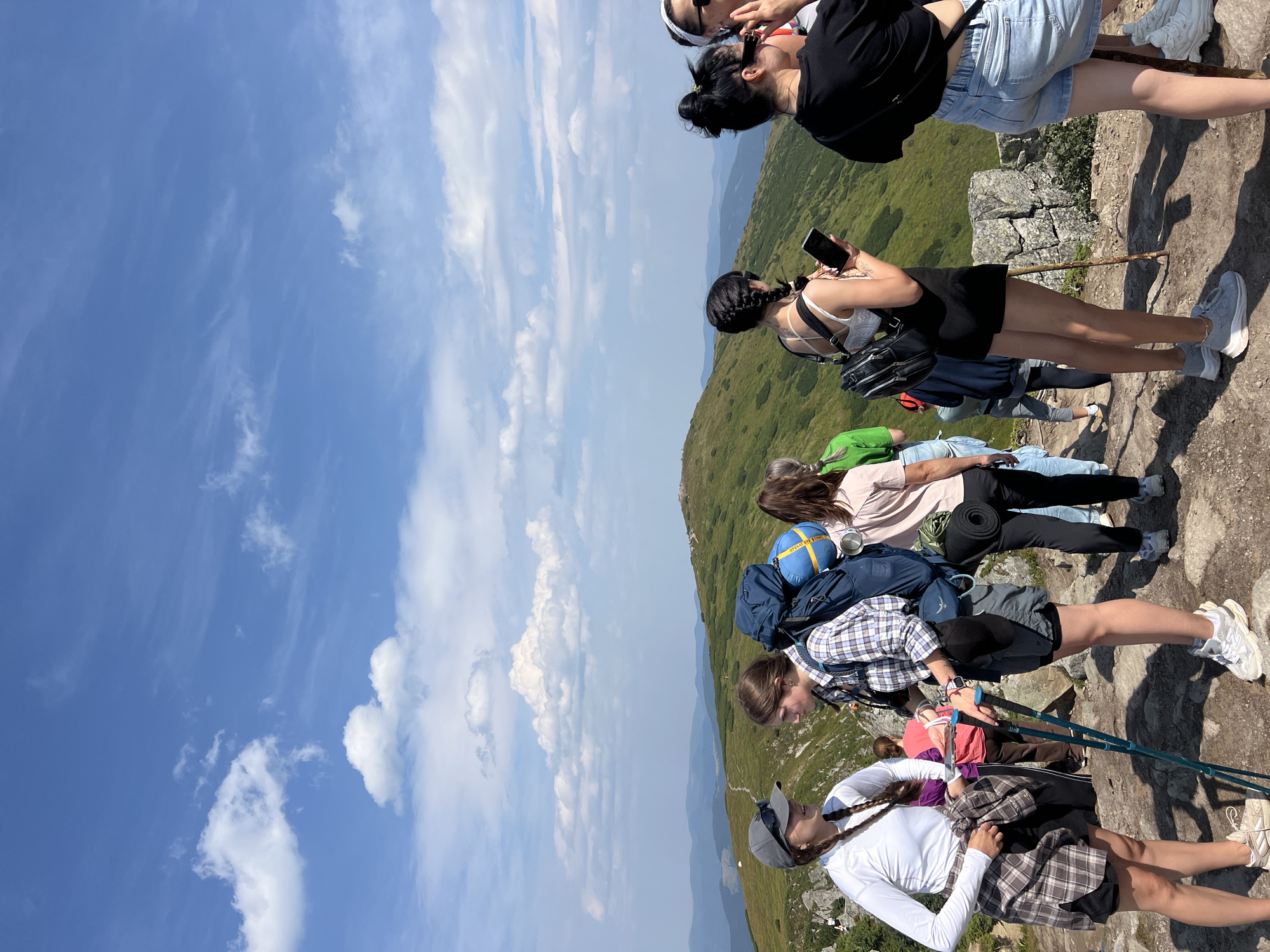 A group of young adults are standing on a rocky mountain trail under a blue sky with scattered clouds. The terrain behind them is covered in green grass and shrubs, with rolling hills extending into the distance. The individuals are dressed in casual, outdoor clothing, and several are carrying backpacks, indicating they are hiking. One person on the left wears a white long-sleeved shirt and a plaid shirt tied around their waist, along with shorts and a cap. Next to them, another person wears a plaid shirt and shorts, carrying a large blue backpack and using hiking poles. To the right, a person in a black top and shorts holds a smartphone, and another person to their right wears a black top and denim shorts. In the background, more people are visible further down the trail. This scene is likely situated in the Carpathian Mountains near Vorokhta, Ukraine, a region known for its mountainous landscape and hiking opportunities.