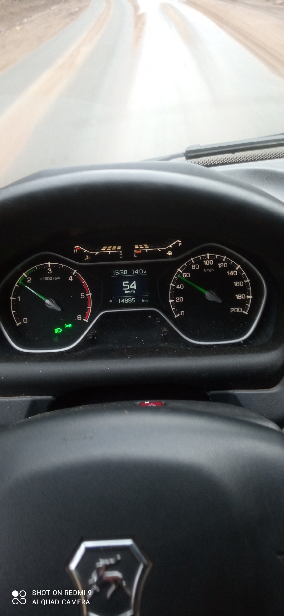 The image depicts the interior of a vehicle traveling on a road. The dashboard indicates a speed of 54 km/h and an odometer reading of 14885 km. The time is 15:38, and the voltage is 14.0V. The low beam headlights are on. The location, as indicated by the logo on the steering wheel and the OCR data, is most likely in Прикамский, Russia. The road ahead appears to be a paved surface with a slight curve.