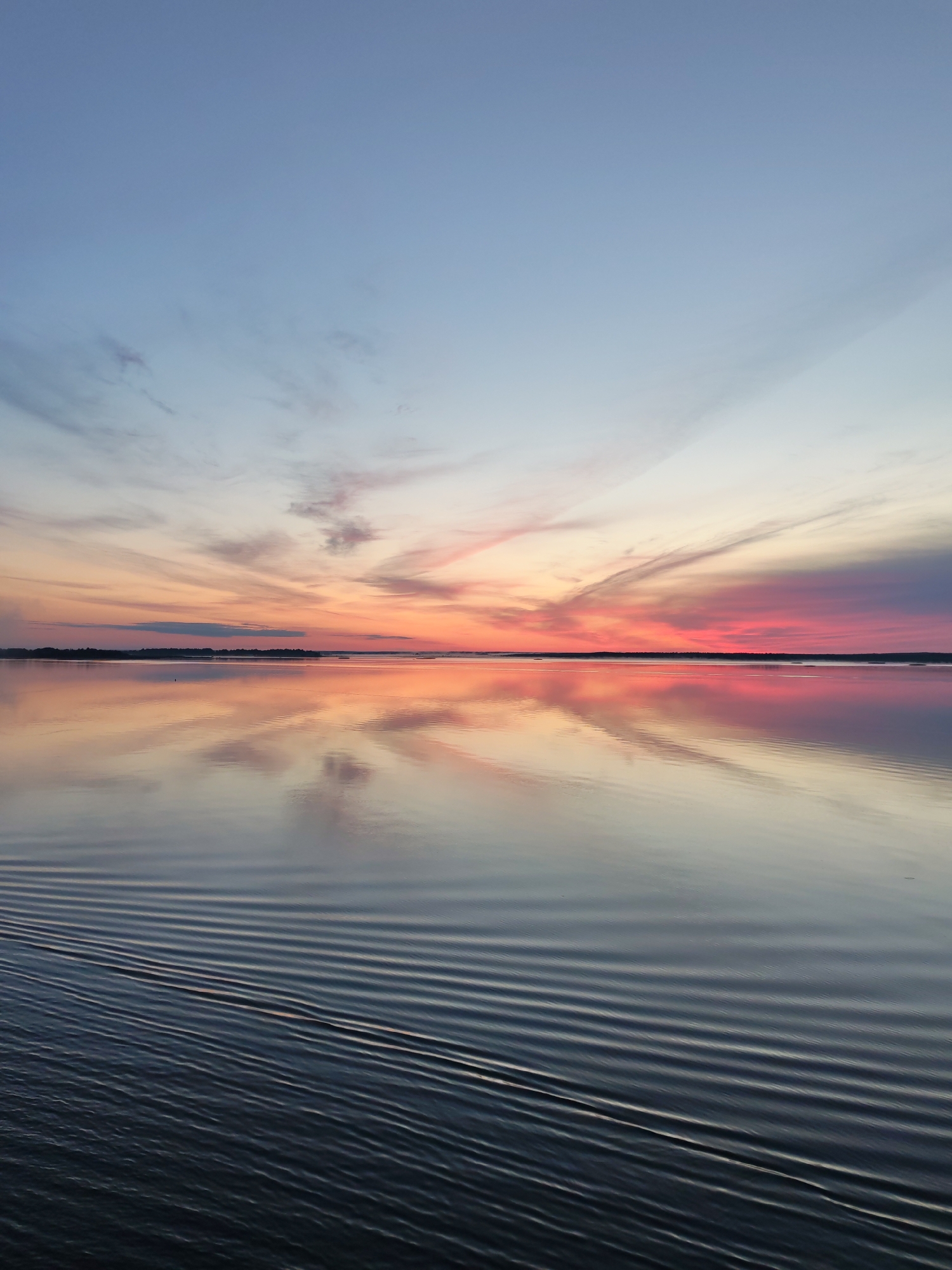 This is a vertical, low-angle, full-shot photograph of a lake at sunset, near Chaika, Russia. The sky displays a gradient of colors, transitioning from a pale blue at the top to vibrant pinks and oranges near the horizon. Wispy clouds are scattered across the sky, catching the sunset's light. The calm water of the lake perfectly reflects the colorful sky, creating a mirror image. In the foreground, the dark water is disturbed by gentle ripples, possibly from a boat. A thin, dark line of trees or land is visible along the horizon. The overall scene is serene and peaceful.