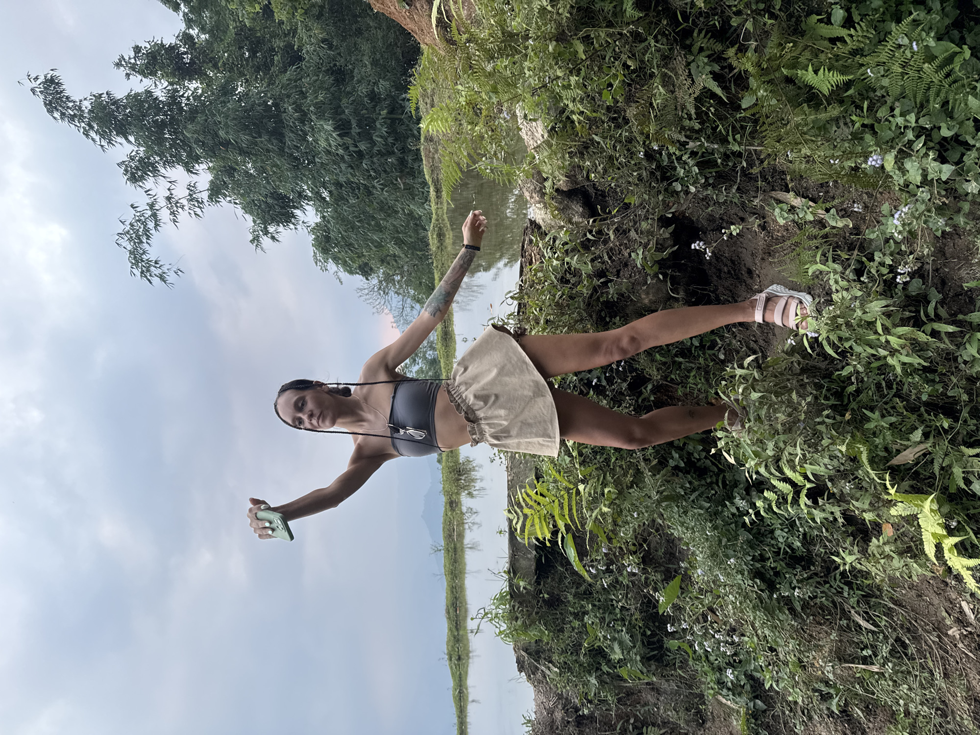 A young woman stands on a lush, vegetated bank beside a body of water, posing for the camera in an outdoor setting. She is positioned towards the right side of the frame, facing left (when the image is correctly oriented). Her dark hair is styled in braids, and she wears a dark gray crop top, a light beige or tan high-waisted skirt, and light-colored sandals. Tattoos are visible on her left arm. In her right hand, she holds a light green smartphone, and sunglasses hang from a necklace around her neck.
The immediate surroundings are covered in dense green foliage, including ferns and various plants with small white flowers, growing from dark soil. To her left, a tranquil body of water, possibly a river or lake, stretches into the midground, reflecting the cloudy sky and the distant landscape. Across the water, the background features more dense green trees or bamboo groves, with faint outlines of mountains or hills beyond.
The scene depicts a serene natural environment under an overcast sky, suggesting a daytime setting with soft, diffused light, possibly morning or late afternoon. The woman appears relaxed, enjoying the scenic view. The location cues, such as the lush vegetation, water, and distant hills, are consistent with Nậm Dịch Commune, Vietnam. No discernible text is visible in the image.