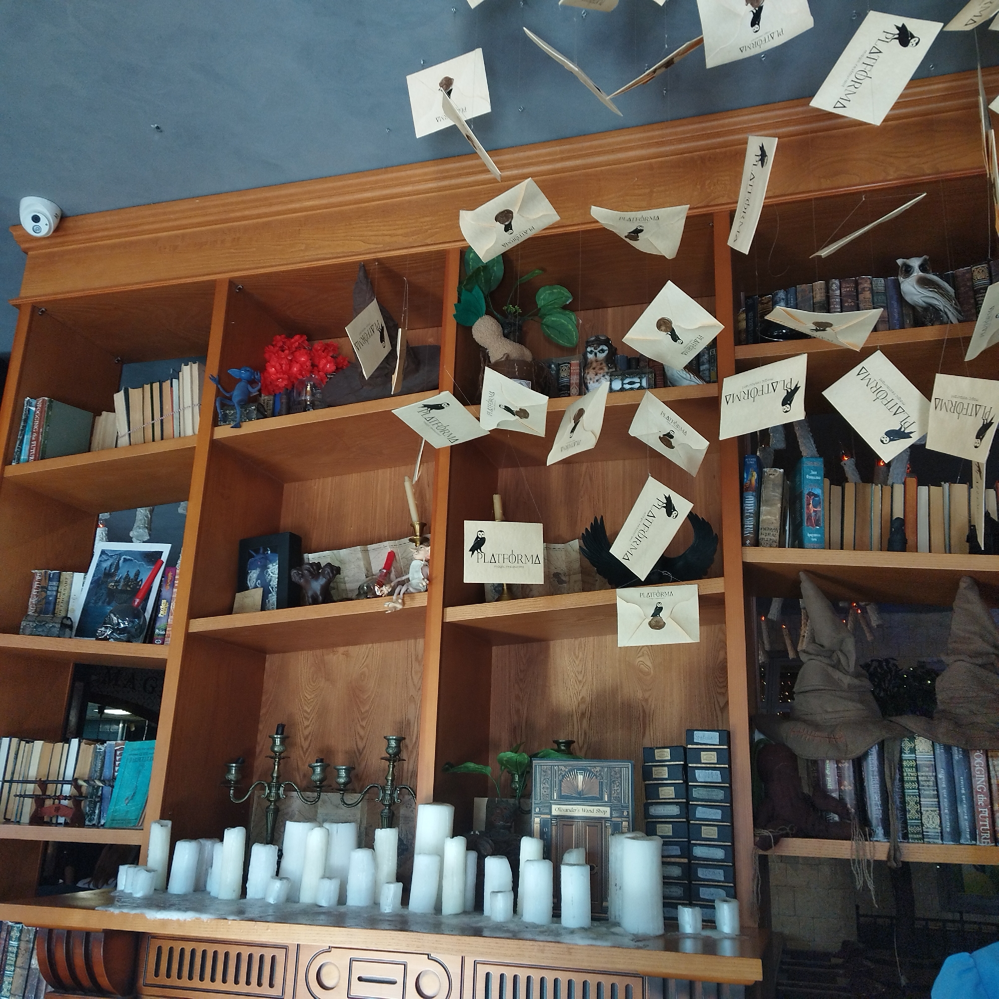 An interior shot reveals a section of a elaborately decorated wooden bookshelf, evoking a Harry Potter theme, likely within a "PLATFORMA magic restaurant" as indicated by recurring text. Numerous cream-colored envelopes, many featuring an owl silhouette and the word "PLATFORMA," are suspended from the light blue/grey ceiling by thin strings, creating the impression of flying letters. A white dome-shaped security camera is mounted on the ceiling in the top left corner.
The shelves are filled with various objects contributing to the magical ambiance. Books, including some with titles like "POGGING the FUTURE," "Waty," and "Wecki," line the shelves. Decorative items include several owl figurines (one prominent white, one smaller brown), a blue creature figurine (possibly a Niffler), a multi-headed dog sculpture, and a wand. Two brown, crumpled hats resembling the Sorting Hat are visible on the far right. A significant detail is a row of white, wax-dripping candles and a classic candelabra on the bottom shelf, adding to the enchanted atmosphere. A box labeled "Ollivander's Wand Shop" is also present on this shelf. Red artificial flowers or berries are arranged in a vase on an upper left shelf. The overall setting is clearly designed to immerse visitors in a fantasy world.