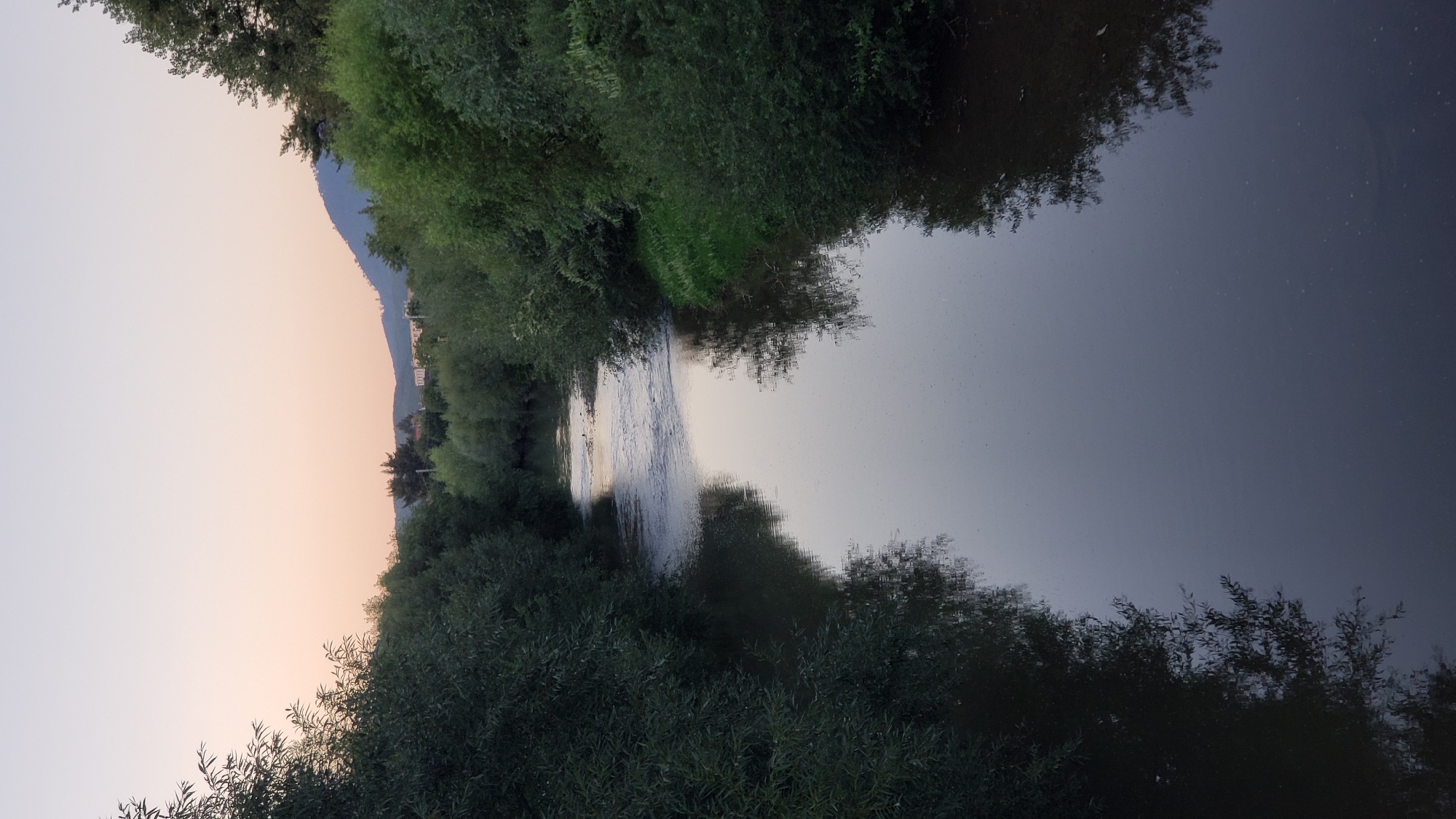 This is a vertical, eye-level shot of a calm river flowing through a densely wooded area at dusk. The river is wide and reflects the soft, pale pink and orange hues of the twilight sky. The water surface is mostly smooth with some ripples visible in the distance, suggesting a gentle current. On either side of the river, lush green trees and bushes line the banks, their foliage forming a thick canopy that overhangs the water. The trees are dense and dark, their reflections casting long, distorted shapes on the water's surface. In the background, the land rises to form rolling hills or mountains, silhouetted against the fading light. A few indistinct buildings and trees are visible on the slopes of the hills. The overall atmosphere is serene and peaceful, characteristic of a natural landscape at the close of day. It is not possible to determine the specific location in Prešov, Slovakia, from this image alone.