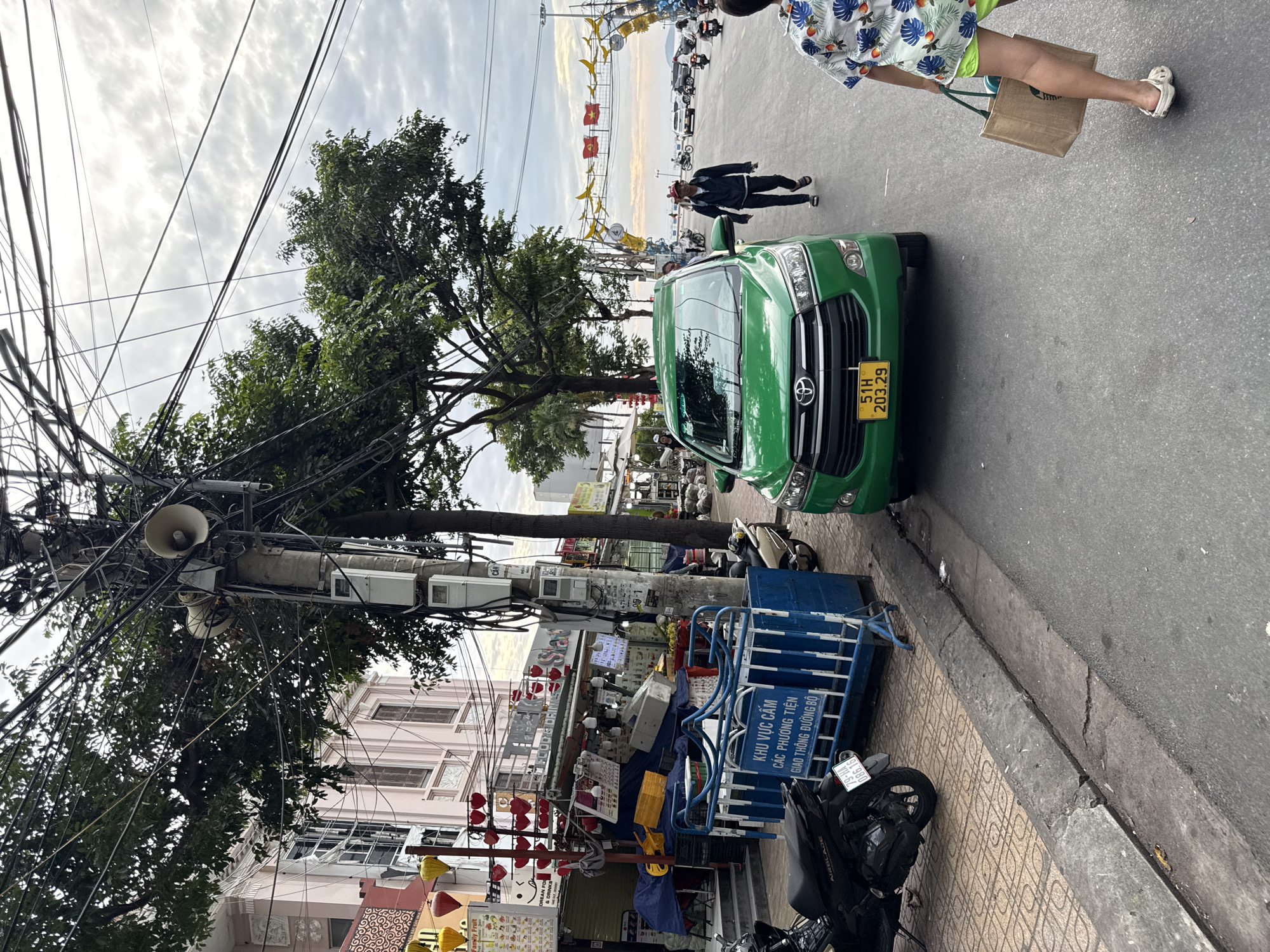 The image depicts a street scene in Nha Trang, Vietnam, likely along a coastal road given the decorative arches with what appear to be flags and banana motifs in the background, suggesting a festive or tourist area. In the foreground, a bright green Toyota Innova taxi is parked on the right side of the asphalt road. A man wearing a dark jacket, dark pants, and a red cap is walking away from the taxi, towards the center of the road. To the right of the taxi, a person wearing a floral print shirt and bright green shorts walks away from the camera, carrying a brown paper bag.
On the left side of the road, a sidewalk is lined with shops and street vendors. Several scooters are parked along the sidewalk. A blue and white metal barrier is placed in front of one of the shops, with Vietnamese text visible on a sign attached to it, indicating a restricted area. The shops appear to sell food and drinks, with various items displayed under canopies. A large utility pole covered in numerous electrical wires and speakers stands prominently on the sidewalk, near the shops. The buildings behind the shops are pale pink, with visible architectural details. The sky is overcast with scattered clouds. The overall scene captures a moment of daily life in a bustling Vietnamese city.