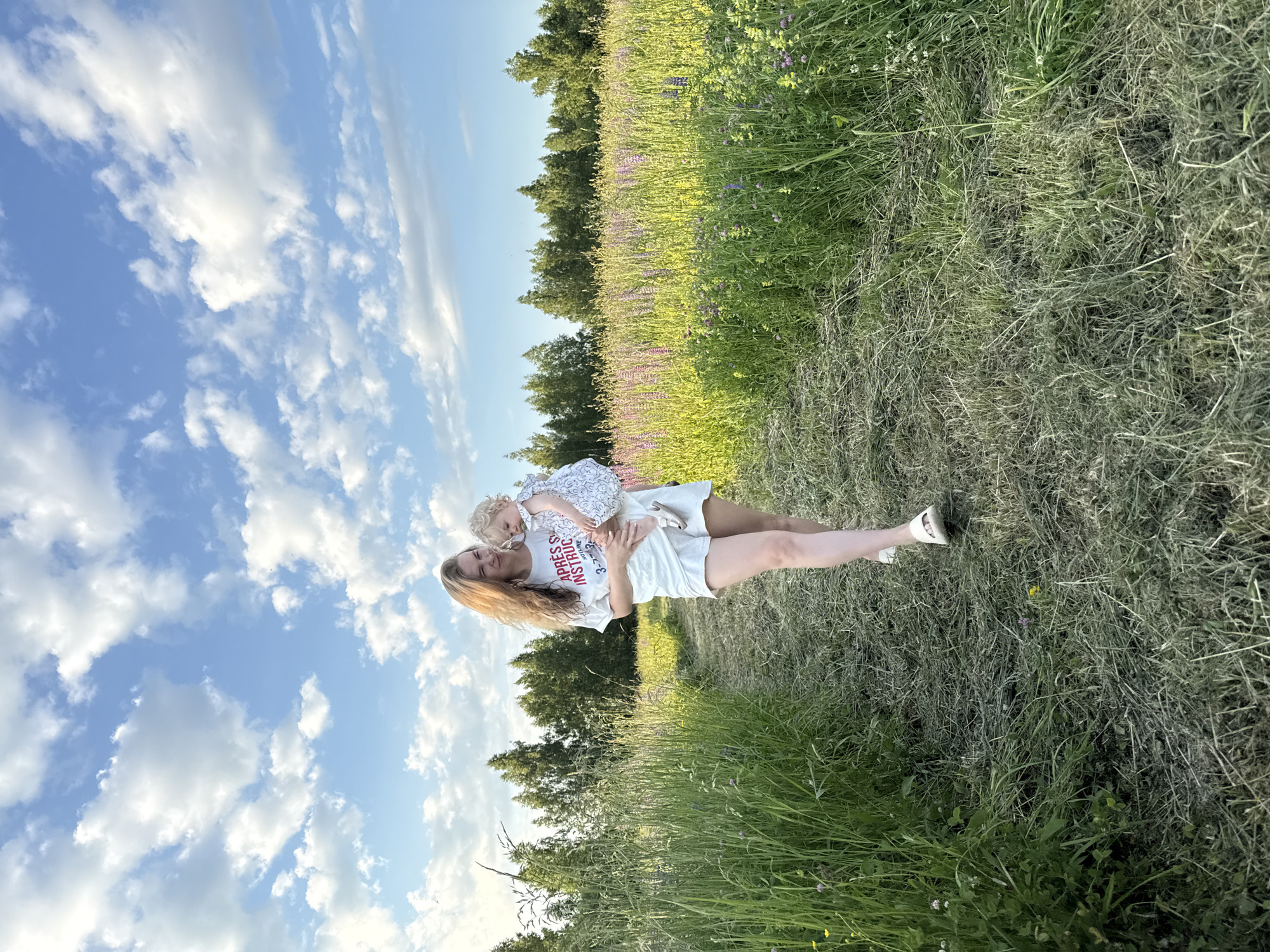 A woman with long, wavy blonde hair is holding a blonde toddler in her arms as they walk along a mown grass path. The woman is wearing a white t-shirt with red lettering that reads "APRES INSTINCT" and white shorts. The toddler is wearing a white and blue patterned dress. They are in a field with tall green grass and purple and yellow wildflowers. In the background, there is a line of trees under a blue sky with scattered white clouds. The scene appears to be in a rural outdoor setting.