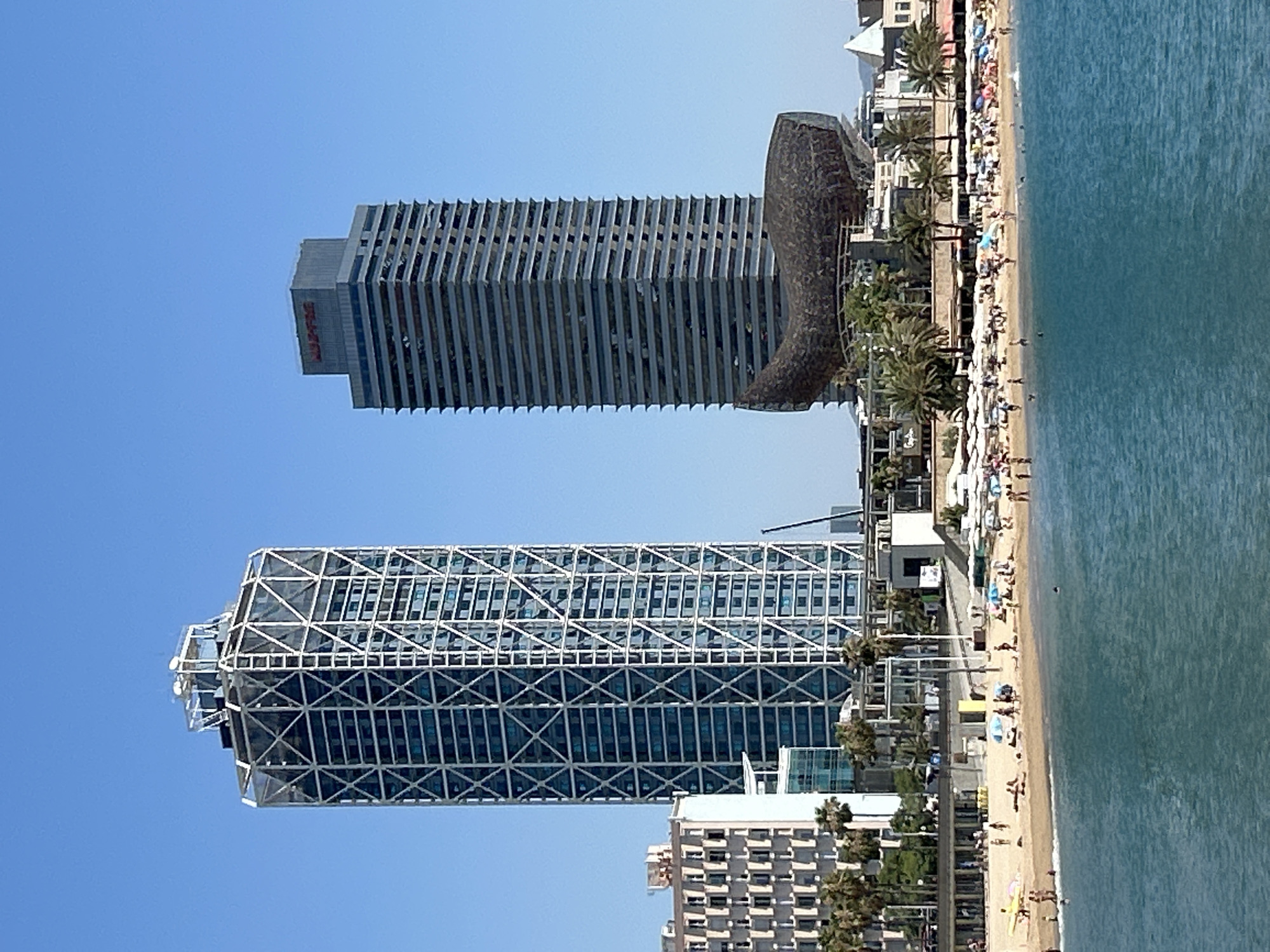 The image shows a view of the beach and skyline of Barcelona, Spain. In the foreground, the turquoise water of the Mediterranean Sea is visible. A sandy beach is populated with numerous beachgoers, many of whom are sunbathing or walking. Beach umbrellas and towels are scattered across the sand. Behind the beach, several buildings are visible. Two prominent skyscrapers stand out: the Hotel Arts, characterized by its distinctive white lattice exoskeleton, and the Mapfre Tower, a more conventional high-rise with horizontal banding. Between the skyscrapers and closer to the beach is a large, dark, fish-shaped sculpture, which is part of the Frank Gehry's "Peix" (Fish) sculpture, located near Port Olímpic. The sky is a clear, vibrant blue, indicating a sunny day. The overall scene depicts a lively urban beach environment.
