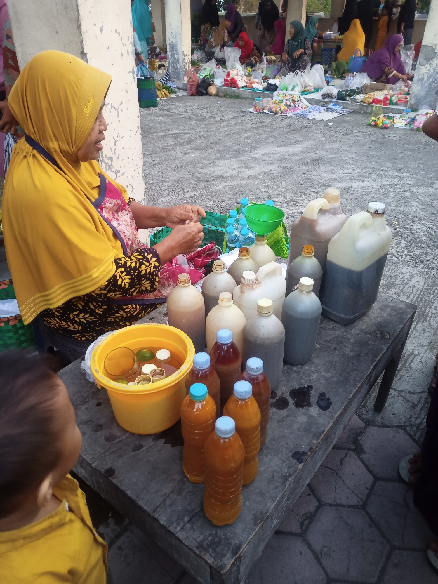 In the city of Nganti, Indonesia, a woman wearing a yellow hijab and a patterned apron is preparing beverages. She sits behind a wooden table laden with various bottles and a yellow bucket filled with liquid and what appear to be citrus fruits. Behind her, a bustling market scene unfolds with other vendors and shoppers. The ground is paved with hexagonal tiles. The overall impression is that of a lively outdoor market with a focus on food and drink.