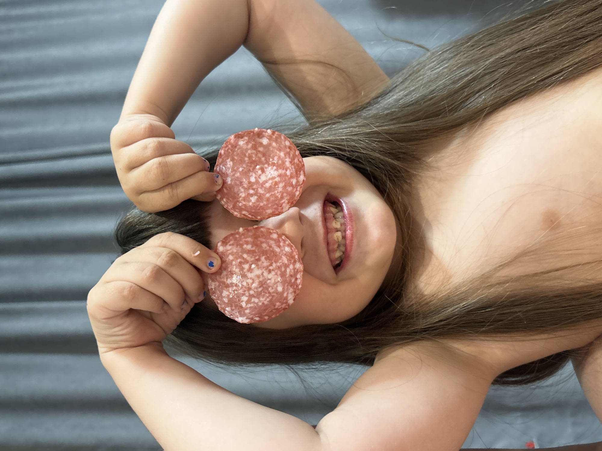 This is a close-up, overhead shot of a young child lying on their back, with their head tilted upwards. The child has long, light brown hair that is spread out around their head. They are holding two round slices of salami over their eyes, with their hands cupped around them. Their mouth is open, revealing their teeth, and their eyes are hidden behind the salami. The child appears to be looking up towards the camera with a playful expression. The background is a muted, textured gray surface, possibly a bed or a mat. The lighting is soft and diffused, suggesting an indoor setting. There is no visible text in the image.