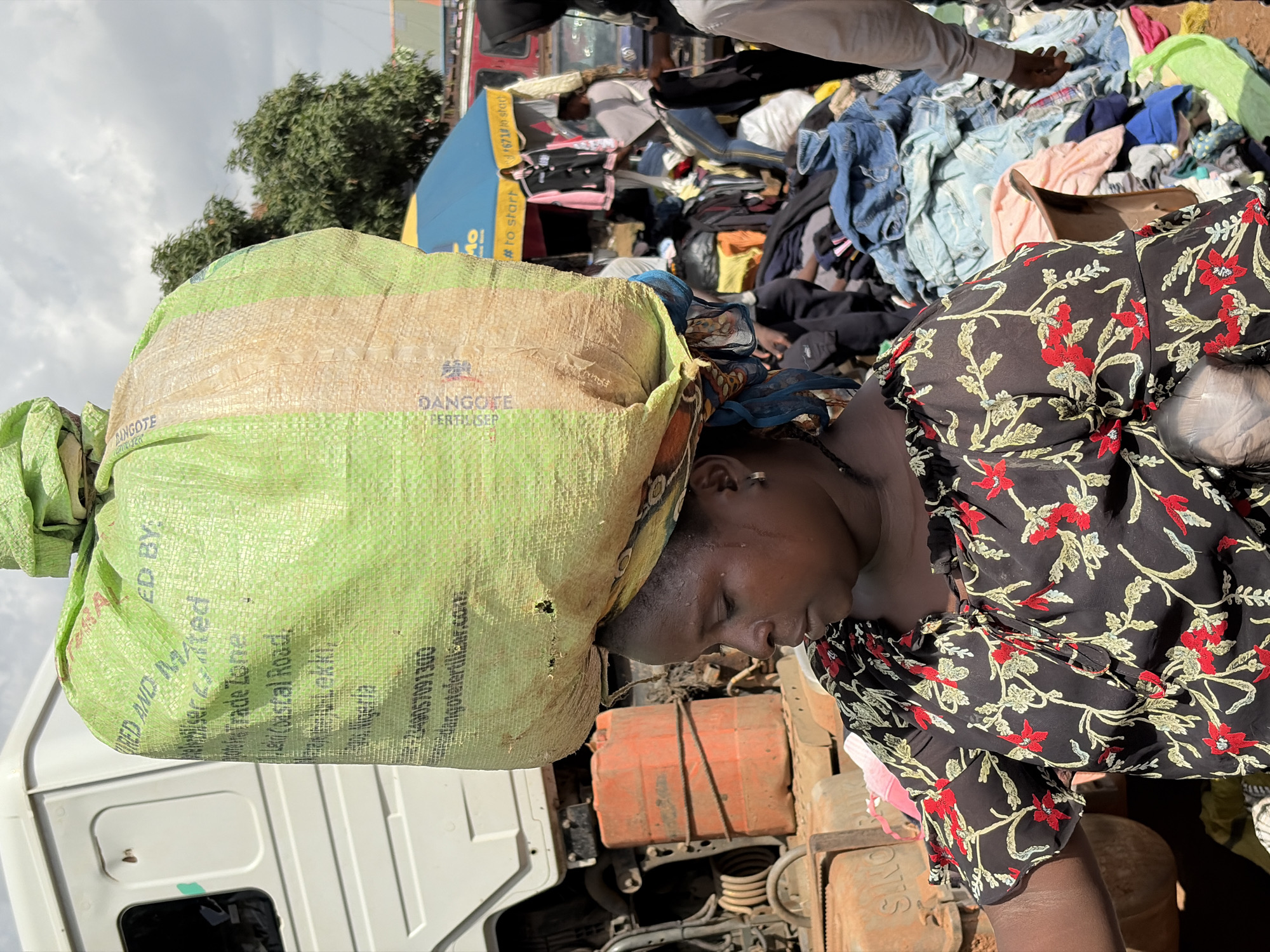 A woman in Gura Top, Nigeria, balances a large, light green sack on her head. She wears a black blouse with a red and yellow floral pattern. The sack appears to contain granular material, possibly fertilizer, as indicated by text on its surface, "DANGOTE FERTILIZER." Behind her, a white truck is visible, along with stacked orange containers. In the background, a busy marketplace unfolds with stalls, umbrellas, and piles of clothing. Other individuals are seen browsing or working in the market. The scene captures a moment of daily commerce in the city.