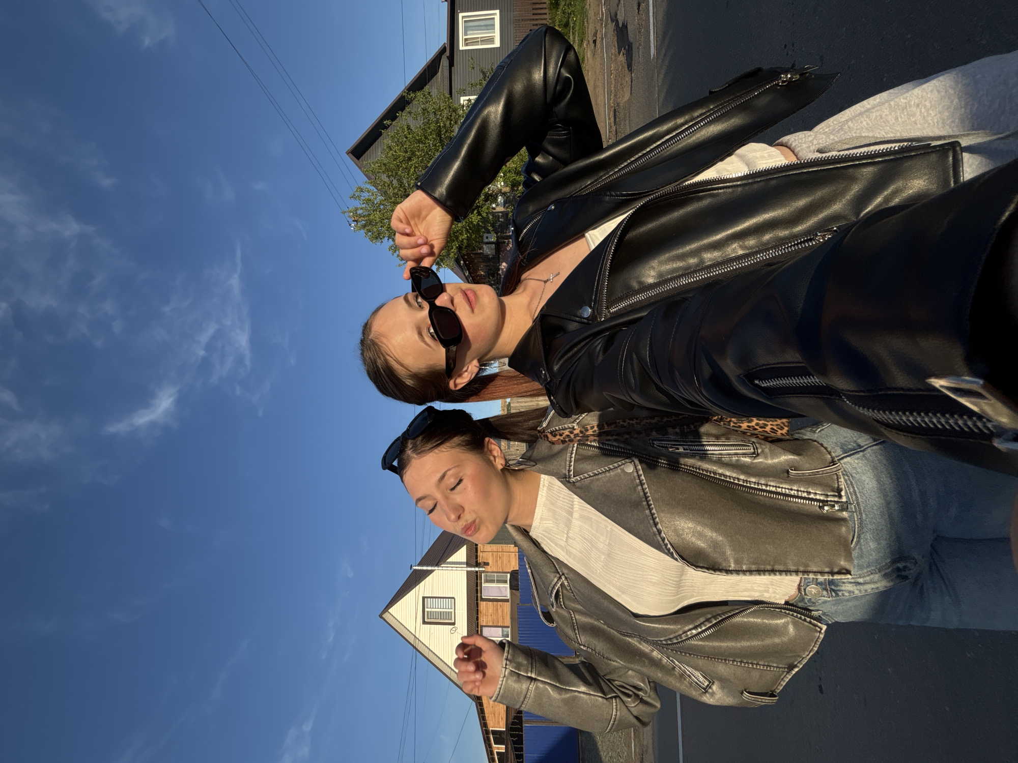 Two young women stand on a street in what appears to be the city of Фоки, Russia, based on architectural styles. The upper woman is wearing a black leather jacket, white top, and sunglasses and is holding her sunglasses near her face. The lower woman wears a distressed gray leather jacket, a white top, jeans, and sunglasses on her head, while she is making a kissing face. Behind them are residential buildings, and a clear blue sky. The perspective of the image is tilted, as though taken from a low angle and rotated.