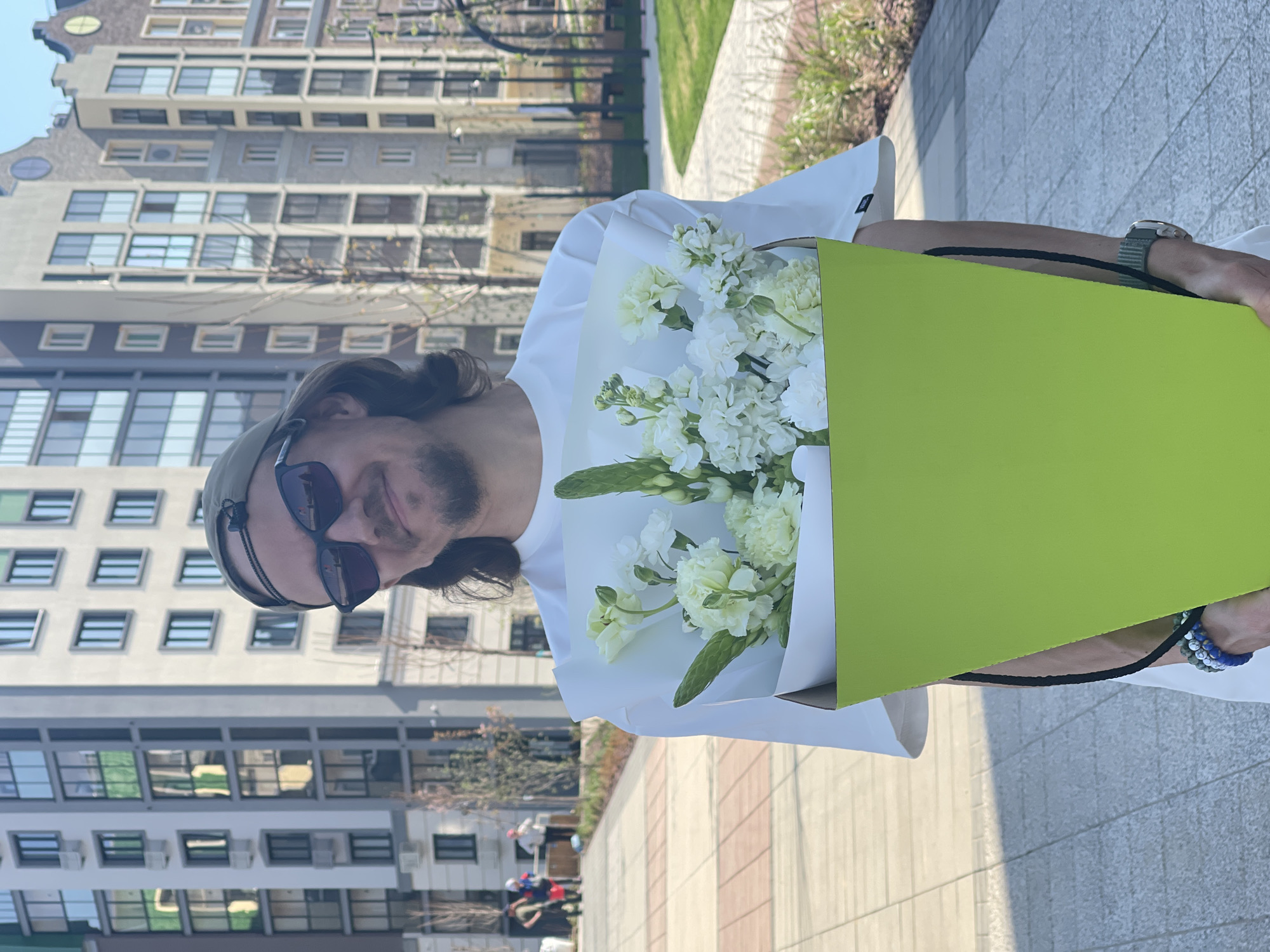 A man wearing sunglasses and a hat is holding a bouquet of white flowers in a green container. He is wearing a white shirt and a watch, and has bracelets on his wrist. He is standing on a paved surface. In the background, there is a building with many windows. The photograph was taken in Хохряки, Russia.