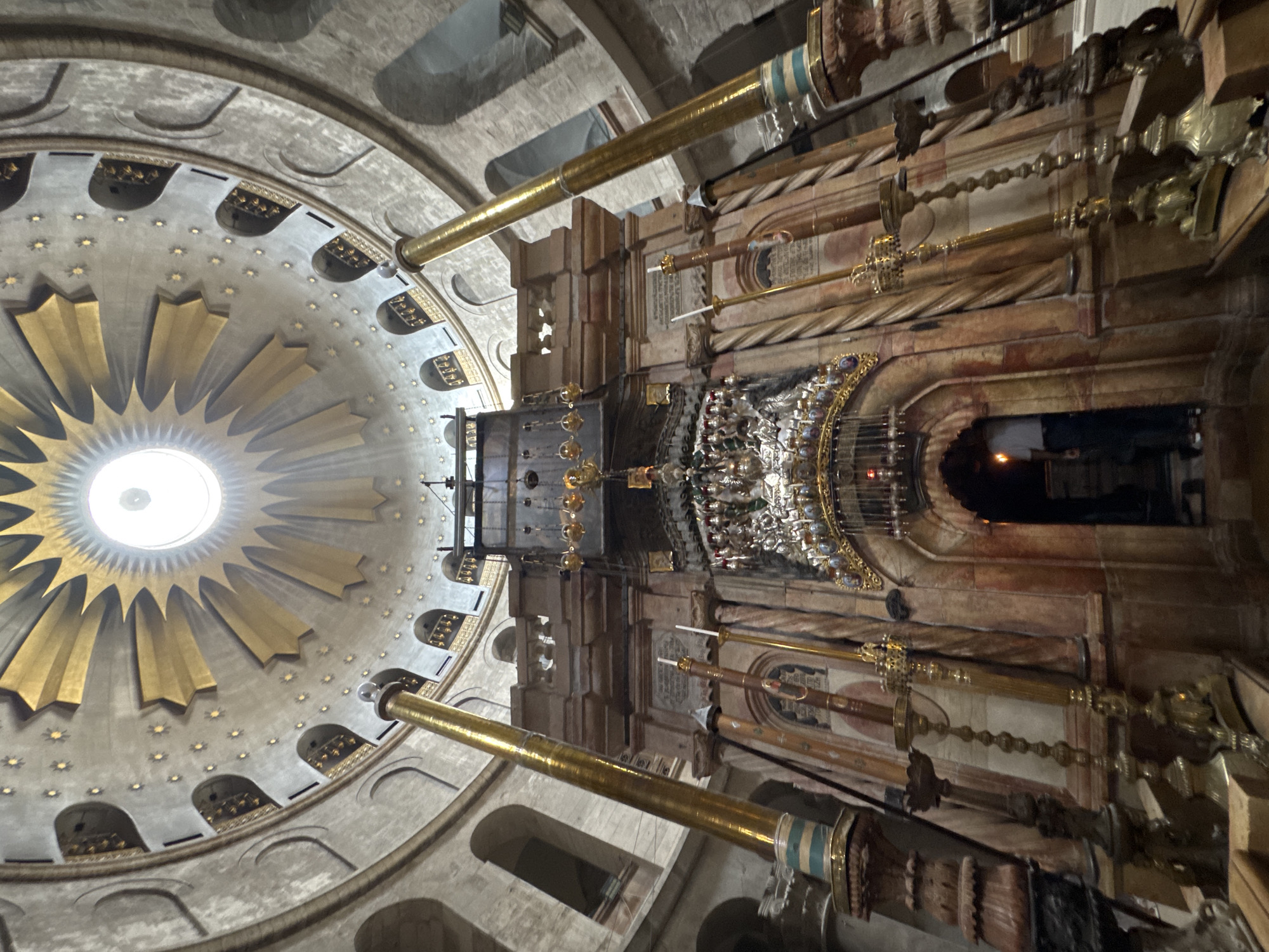 The image depicts the interior of the Holy Sepulchre in Jerusalem, Israel. The focal point is the Aedicule, a structure built over what is traditionally believed to be the tomb of Jesus. The Aedicule is elaborately decorated with marble and ornate metalwork, including lamps and chandeliers. Above the Aedicule, a large, domed rotunda features a circular opening at its apex, allowing natural light to filter in. The dome is adorned with gold accents and star-like embellishments, contributing to the grandeur of the space. Several large, golden columns flank the Aedicule. In the background, architectural elements of the church, such as arched windows and stone walls, are visible. A person is partially visible entering an arched doorway beneath the Aedicule. The scene is well-lit, with a combination of natural and artificial light sources.