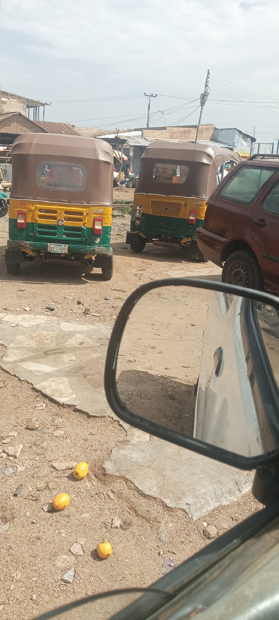 The image shows a street scene in Bukuru, Nigeria. Two yellow and green auto rickshaws (tricycles), commonly known as "keke napep," are parked in the street. A maroon sedan is partially visible on the right side of the frame. Three yellow fruits, possibly mangoes, lie on the ground in the foreground. The view is taken from inside a vehicle, as a side mirror is prominently featured. Background buildings are simple, likely housing local businesses or residences, and are in various states of construction. The sky is overcast. There are electric lines and poles in the background. The ground is a mixture of dirt and concrete.