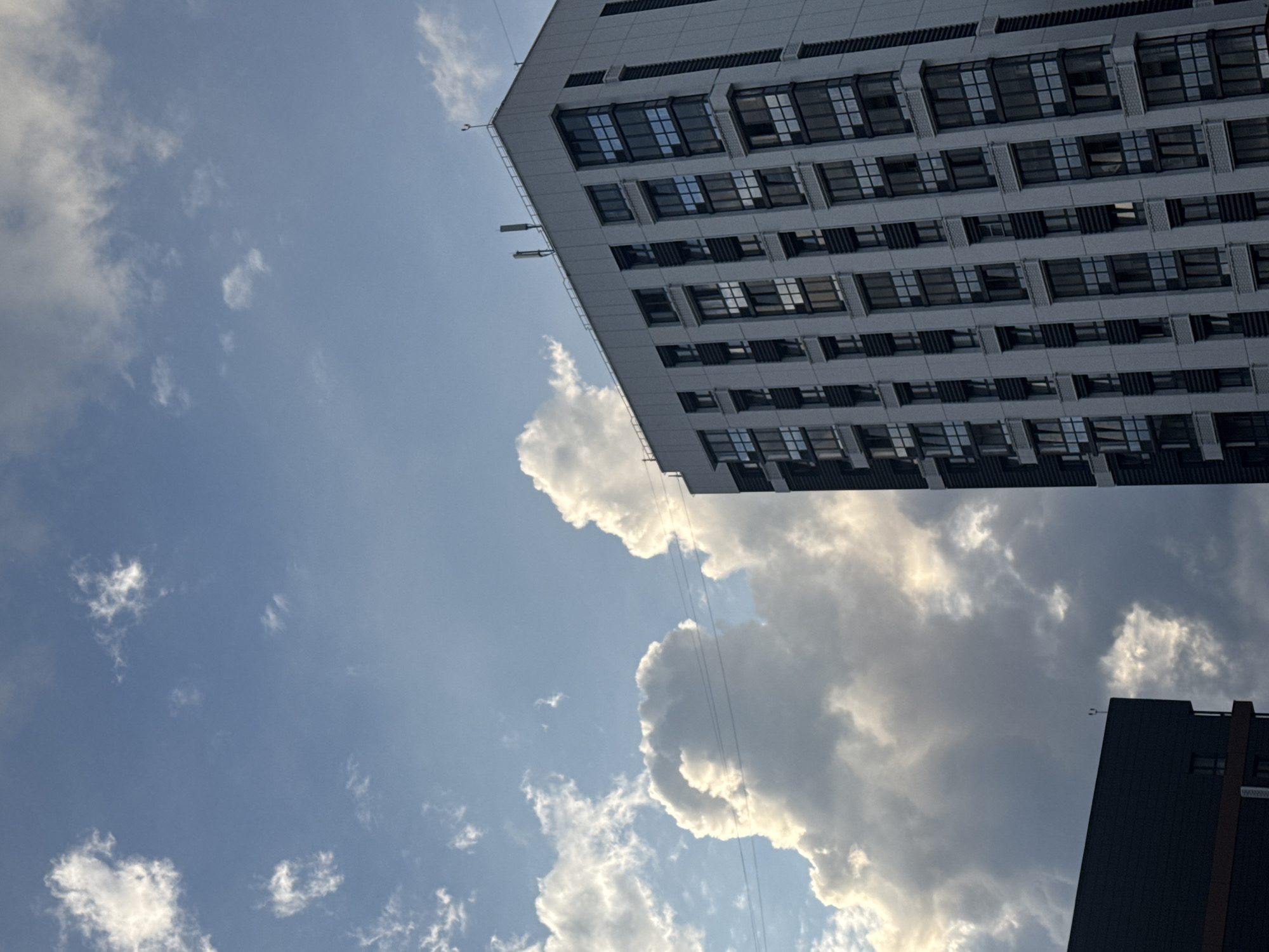 The image shows a modern, tall residential building with many windows, viewed from below against a cloudy sky. The building appears to be a light gray color with dark gray accents around the windows and vertical elements on the facade. Two antennas are visible on the rooftop. In the foreground, the corner of another dark-colored building is visible. The sky is a mix of blue and white, with large, dramatic clouds that are partially illuminated by sunlight. There are power lines stretching across the frame. The scene is likely an urban environment. Based on the architectural style and the presence of similar structures in online image searches, this building is likely located in the city of Novosibirsk, Russia. There are no people visible in the image.