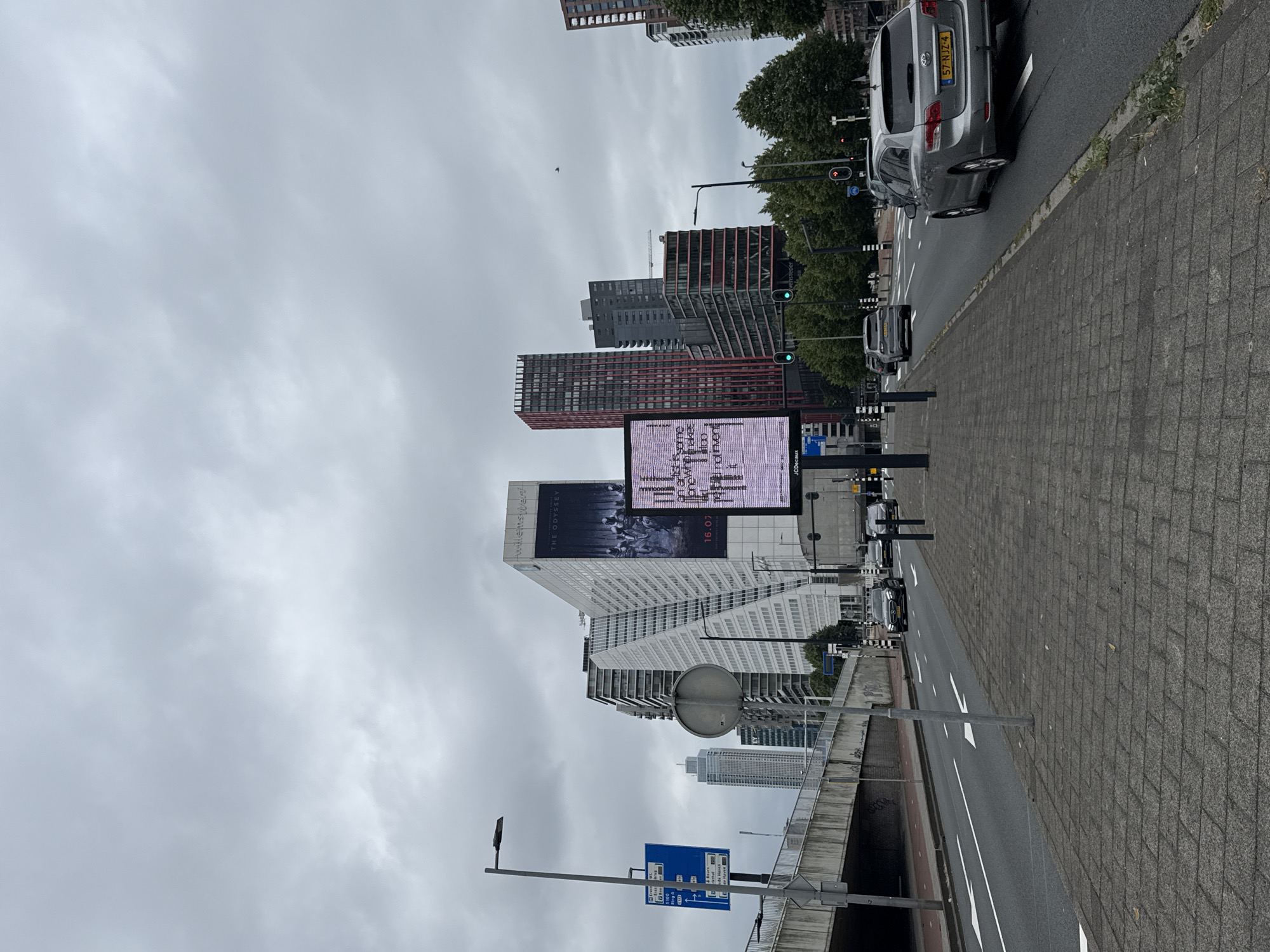 The image shows a street scene in Rotterdam, Netherlands, on a cloudy day. In the foreground, a grey paved sidewalk runs alongside a multi-lane road with traffic. Several cars are visible on the road, including a silver Toyota estate car with license plate "57-NJZ-4". On the left side of the road, there is a pedestrian overpass and a blue street sign with multiple destinations. In the background, a collection of modern buildings rises against the overcast sky, featuring a prominent white building with an angled facade and a large billboard displaying "THE ODYSSEY" and "16.07". To the right, a large digital screen displays scrolling text. Trees line the side of the road further down, and traffic lights are visible. The overall impression is of an urban environment with significant architectural structures and active traffic.