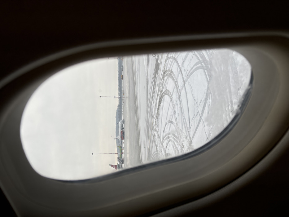 The scene depicts an airport apron viewed from inside an aircraft through an oval window, likely in Novogorelovo, Russia. The external environment is overcast with a muted sky, characteristic of a winter day. The ground outside is partially covered in snow, exhibiting numerous dark tire tracks from ground vehicles, particularly in the foreground and midground areas. Patches of dark, wet asphalt are also visible.
In the midground, two commercial passenger aircraft are present. To the left, a white aircraft with a prominent red tail fin and a green logo (S7 Airlines livery) is stationary, connected to a jet bridge. Ground support equipment, including a set of airstairs and other vehicles, are positioned near this aircraft. To the right of the S7 aircraft, a second white aircraft with a blue and red tail logo (likely Rossiya Airlines, part of Aeroflot Group) is on the tarmac. A red ground service vehicle, possibly a tug or de-icing truck, is positioned near the nose of this aircraft.
Multiple tall, dark grey light poles with arrays of luminaires stand across the apron. In the far background, a dark line of trees or forest is visible under the hazy sky, marking the perimeter of the airfield. No individual people are distinctly identifiable, but their presence is indicated by the operational aircraft and ground vehicles. The overall environment suggests an active airport during cold weather conditions.