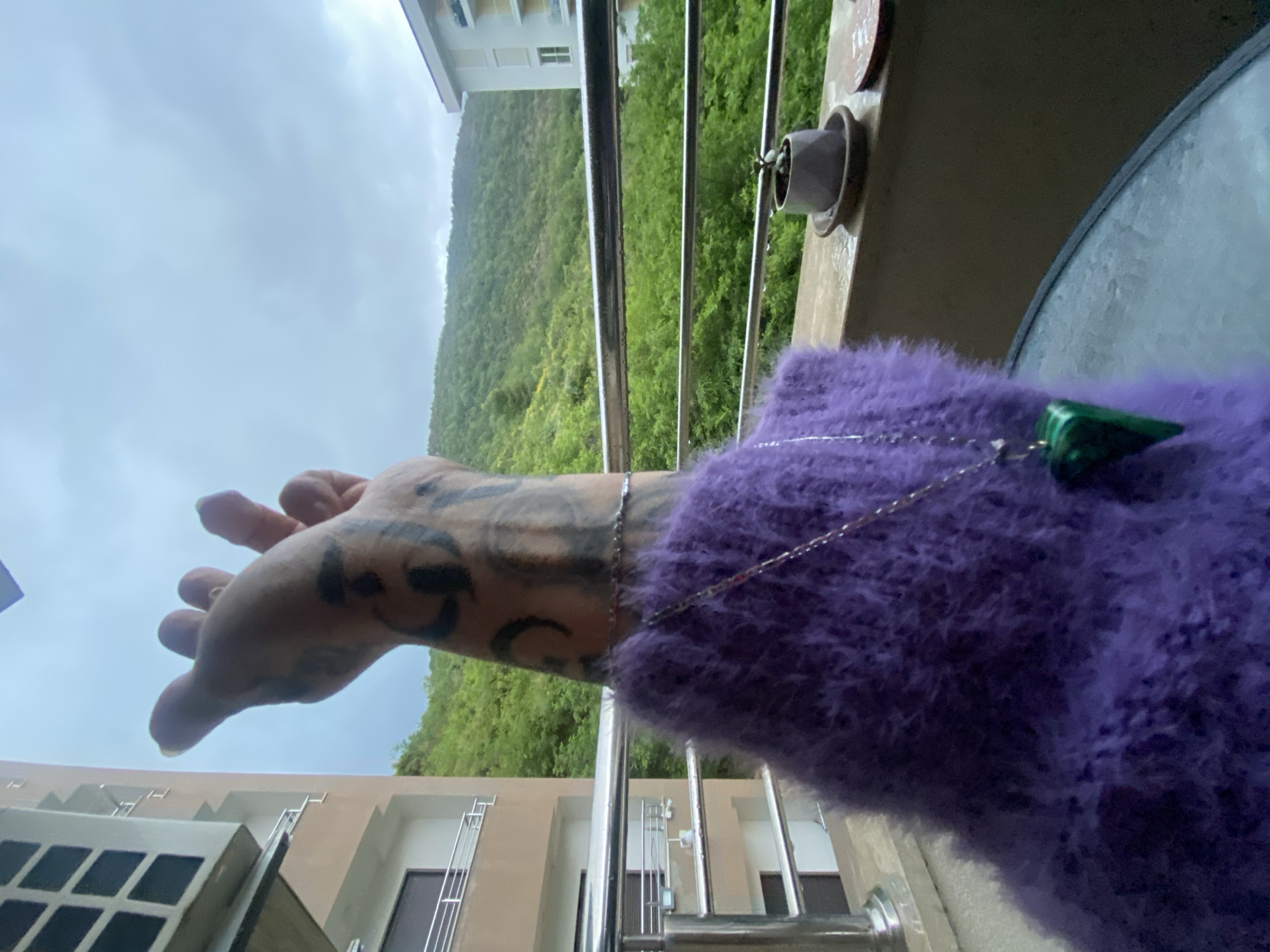 The image shows a person's hand and arm extending over a balcony railing. The hand has tattoos and is positioned with the fingers extended. The person is wearing a purple, fuzzy sweater, and a silver necklace with a green, triangular pendant. The background reveals a view of lush green hills, buildings, and a cloudy sky. The photograph appears to be taken in Boreti, Montenegro.