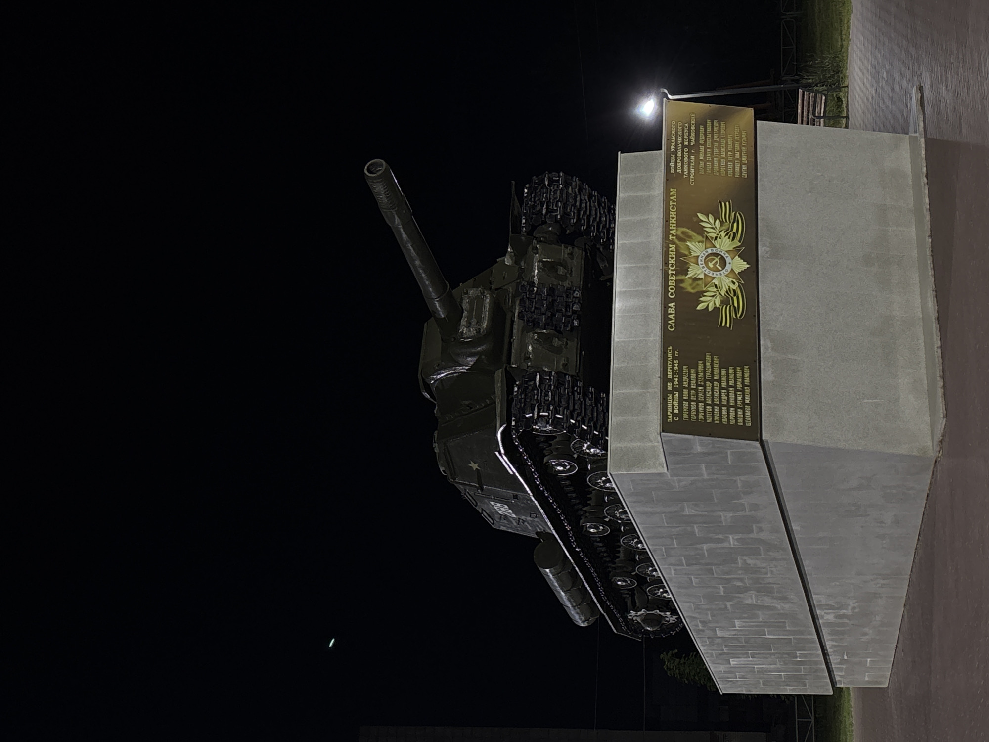 The image shows a monument featuring a Soviet-era self-propelled gun, likely an ISU-152 based on its appearance, positioned on a concrete pedestal. The monument is illuminated at night, with a dark sky in the background. A plaque on the pedestal displays text in Russian and an emblem with a star and laurel wreath. The text on the plaque appears to list names and dates, possibly commemorating fallen soldiers or tank crews. The environment suggests an outdoor public space, with a streetlamp visible to the right and hints of a park-like setting with a bench and some greenery in the distance. The monument is located in the city of Chaikovsky, Russia, as indicated by the inscription on the plaque, "СТРОИТЕЛЯМ г. ЧАЙКОВСКИЙ" which translates to "To the builders of the city of Chaikovsky."