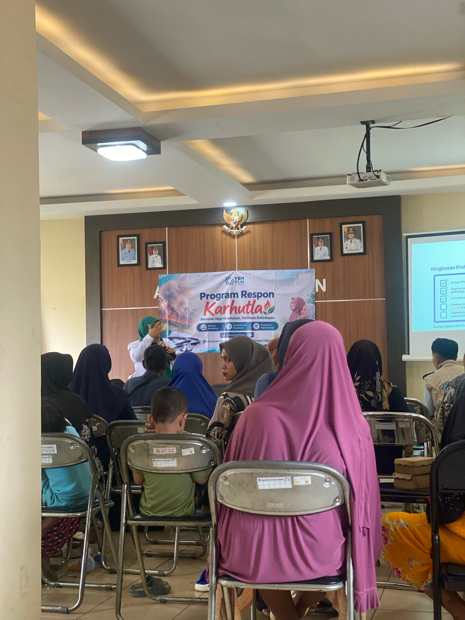 In a room with a projector and a screen displaying "Ringkasan Proyek", a woman in a green hijab gestures while speaking to an audience seated on chairs. The audience consists of men, women, and children, many of whom are wearing hijabs. A banner in the background reads "Program Respon Karhutla" with an image of smoke and fire. The room appears to be a community hall or meeting room in Banjarbaru, Indonesia, indicated by the presence of Indonesian government emblems and text on the chairs and banner. The overall atmosphere suggests an educational or informational session.