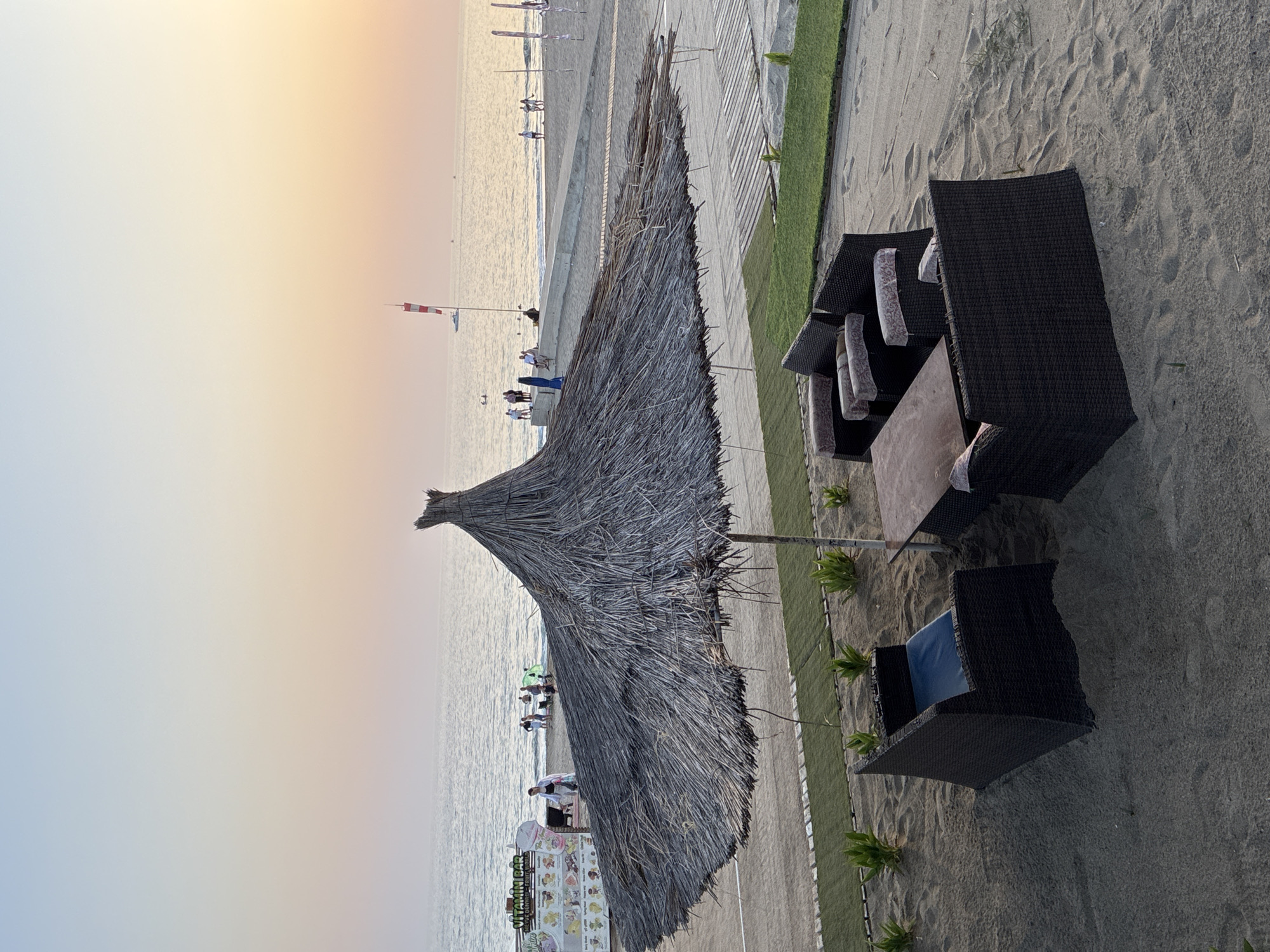 The image depicts a tranquil beach scene in Alanya, Turkey, during the late afternoon or early evening. The foreground is dominated by a large, thatched umbrella providing shade over a seating area situated on the sandy beach. The seating consists of dark wicker furniture, including sofas and a table, with patterned cushions on the sofas. Small green plants are visible in cultivated patches around the seating area.
Beyond the seating area, a paved walkway leads towards the sea. Several people are scattered along the beach and the walkway, some walking, others sitting and relaxing. In the background, the calm sea stretches to the horizon under a gradient sky of soft blues and oranges, indicative of a sunset. A small white boat with a red and white flag is visible on the water, and further out, there appear to be other distant boats or buoys. On the left side of the image, a stall labeled "VITAMIN BAR" with colorful images of fruits and ice cream is set up, suggesting a commercial area within the beach. The overall atmosphere is one of peaceful relaxation by the sea.
