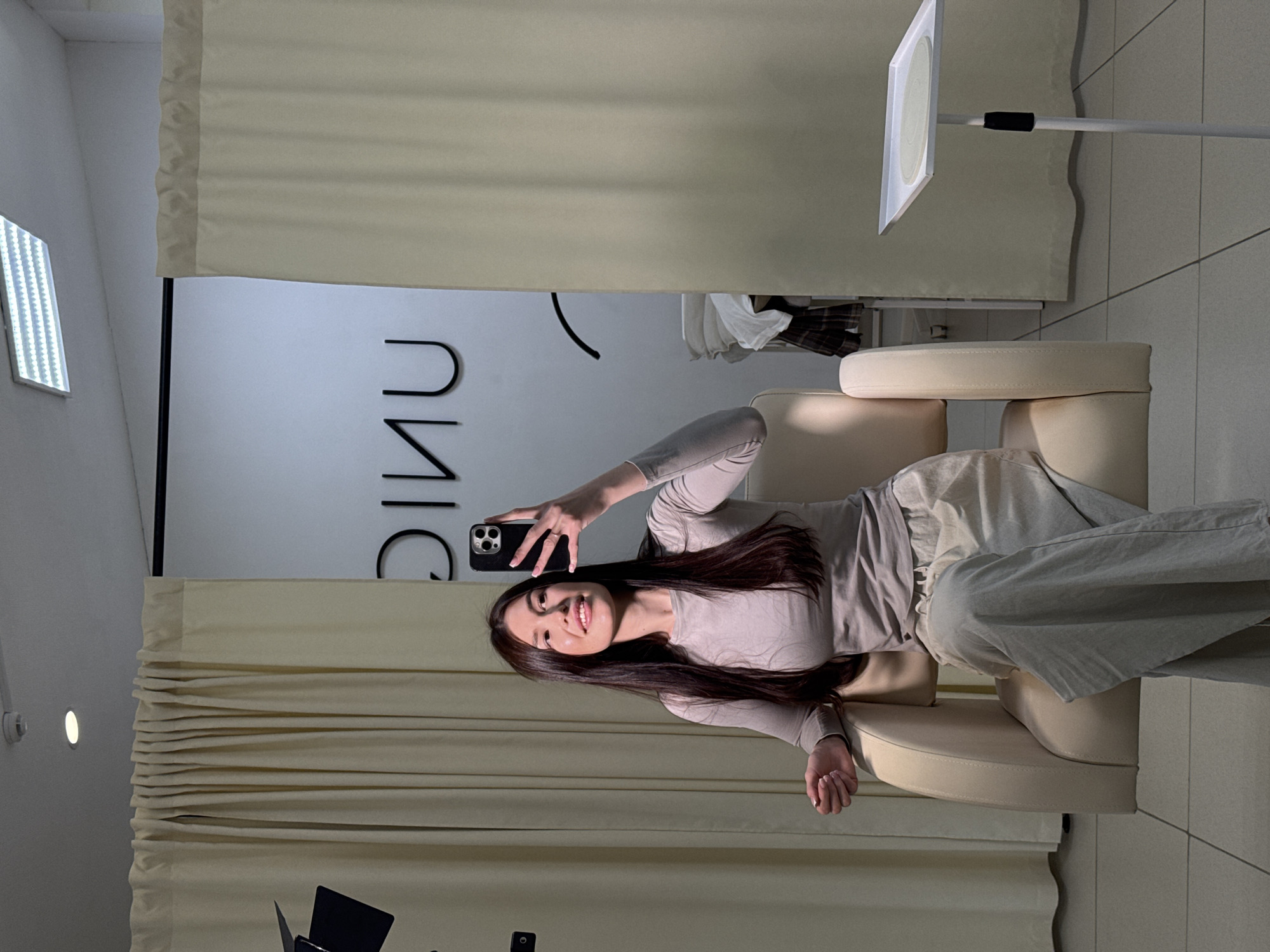 A young woman with long, dark hair is captured taking a selfie in an indoor setting. She is seated in a light beige armchair, wearing a light-colored, long-sleeved top and matching wide-leg pants. Her right hand holds up a black smartphone to take the picture, and she is smiling gently at the camera.
The background features light-colored curtains on either side, framing a light-colored wall. On this wall, large, dark letters are visible, appearing as "UNI" (possibly part of "UNIC" or similar, partially obscured or reversed as if seen in a mirror reflection). To the far left, a rectangular LED light panel is affixed to a white wall. Another light source, possibly a softbox or ring light on a stand, is visible in the upper right portion of the image. The environment appears clean and well-lit, suggesting a modern studio, fitting room, or lounge area in Chaikovsky, Russia. The woman's expression conveys a relaxed and happy mood.