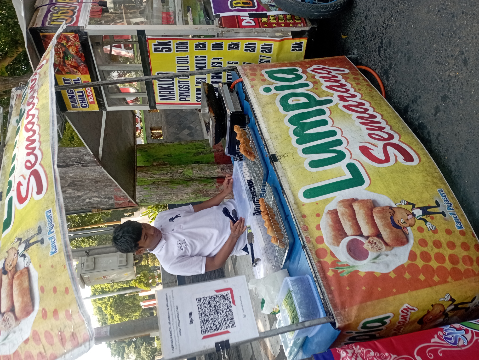 A young man, likely an employee, stands behind a food cart in Nganjuk, Indonesia, preparing food. The cart is decorated with colorful banners advertising "Lumpia Semarang" and other food items. The man wears a white polo shirt with a black horse emblem. He holds a pair of tongs, and in front of him, a metal rack holds several fried spring rolls. To his left, a container holds chopped green vegetables, and to the right, a gas stove with a frying pan is visible. A QR code for payment is displayed on a sign next to the cart. The background shows trees and a utility box. The ground is paved with asphalt.
