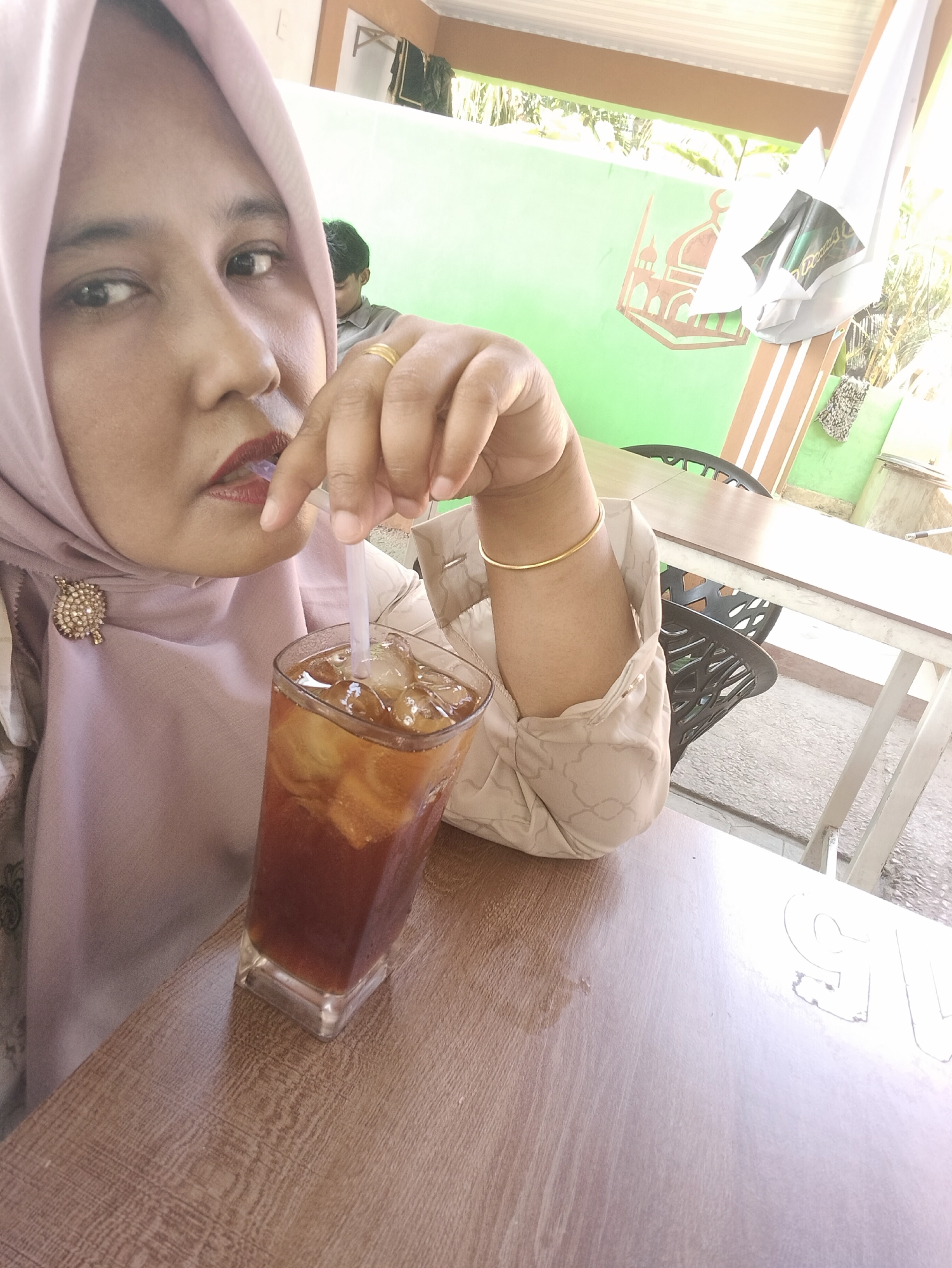 The image depicts a person wearing a pink hijab and a patterned beige blouse, seated at a table and drinking from a glass of iced tea with a straw. A gold-colored bracelet adorns their wrist, and a ring is visible on their finger. In the background, another person is partially visible, seated in the distance. The setting appears to be outdoors, with a green wall decorated with an Islamic-themed design visible. The table is wooden, and the ground is concrete. Based on the location metadata, this scene takes place in Matang, Indonesia.