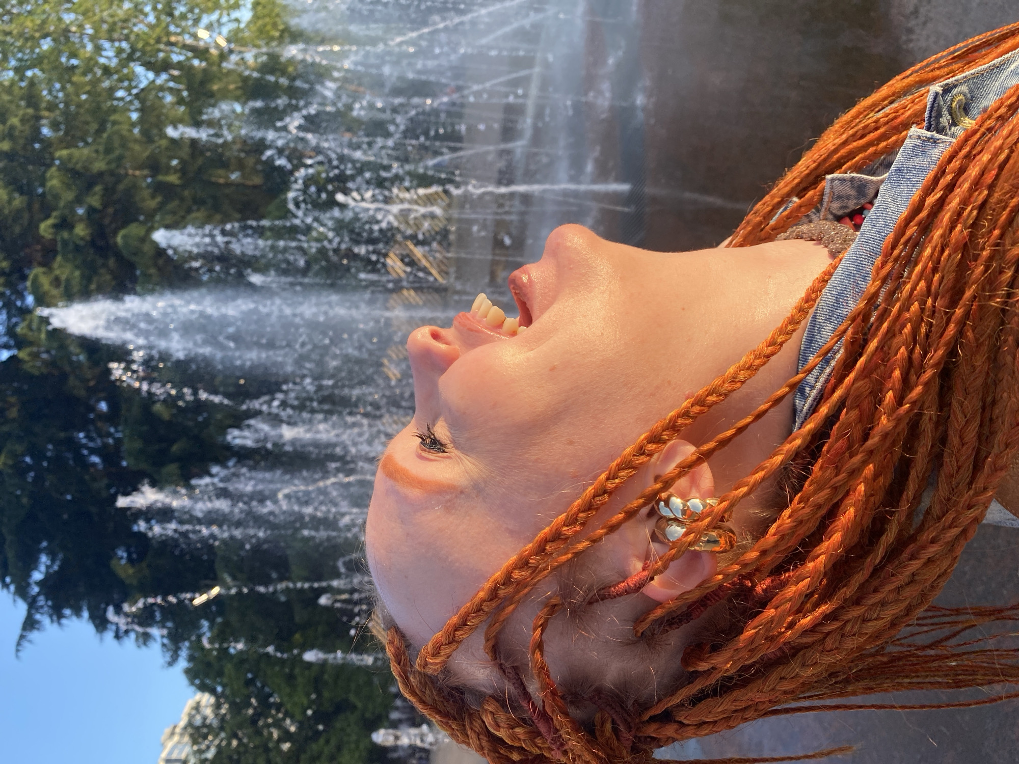 A close-up, profile view shows a person with vibrant orange braided hair laughing with their head tilted back towards a fountain with multiple water jets in Sochi, Russia. The sun illuminates the person's face and hair with a warm glow. The person is wearing a denim jacket over a gray collared shirt. They have a large gold hoop earring in their visible ear. The background is blurred, with the water from the fountain and greenery from trees.