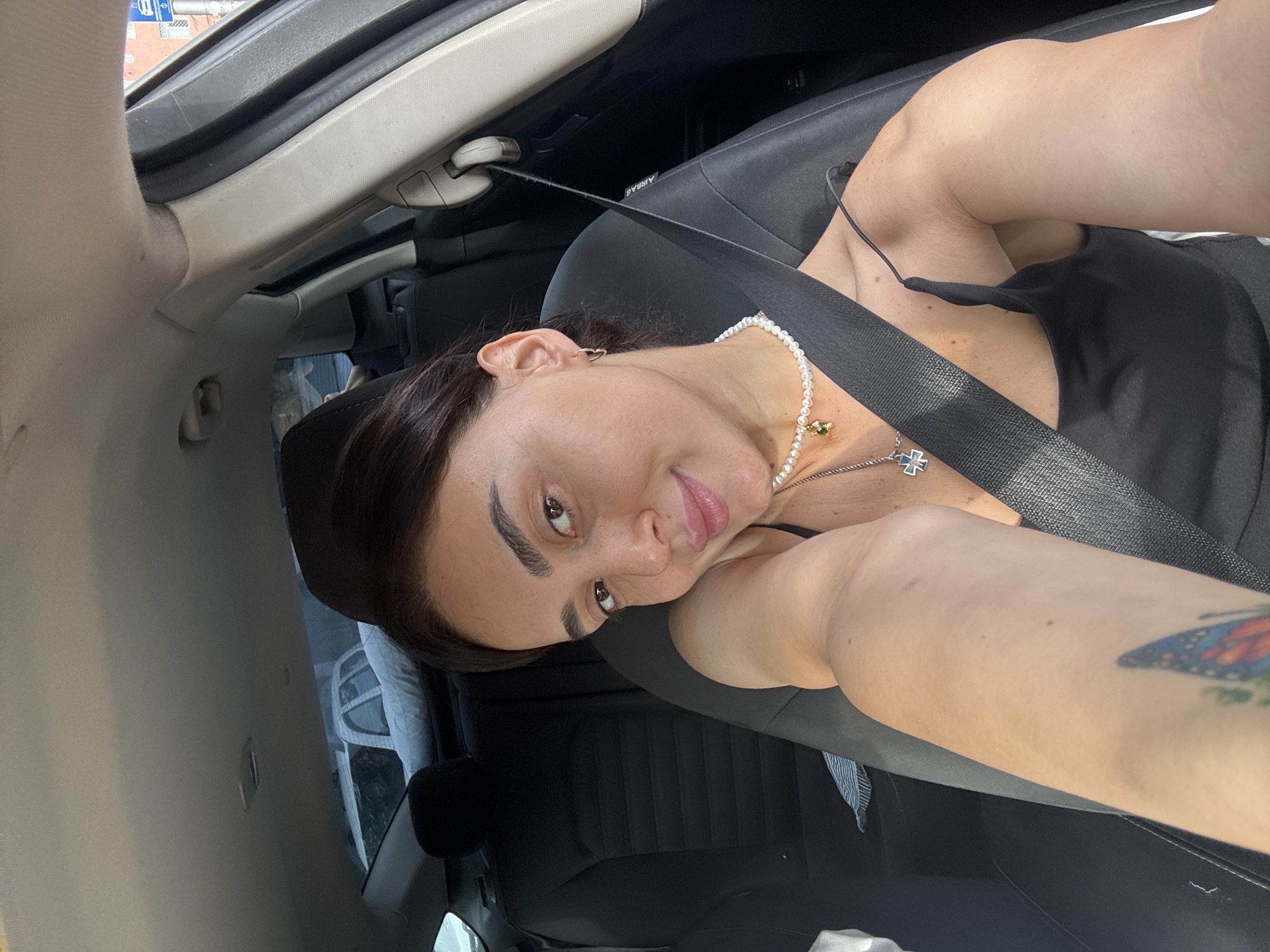 The image shows a young woman with dark hair and brown eyes sitting in the passenger seat of a car. She is wearing a black tank top and a pearl necklace with a pendant. Her left arm is resting on her shoulder. The car interior is visible, including the black seats and the seatbelt. The background shows a street scene with buildings, suggesting the location is in Tbilisi, Georgia. The lighting is natural and appears to be daytime.