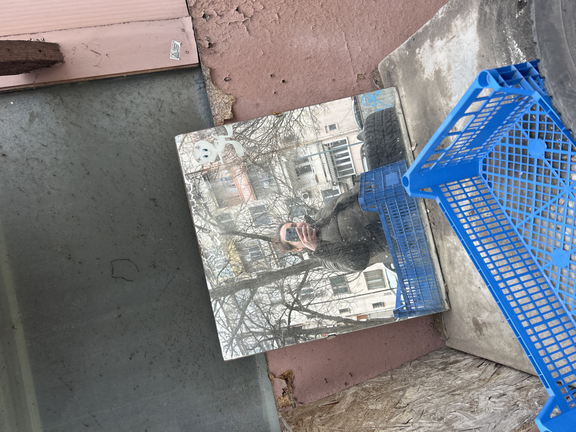 The image shows an outdoor scene in Plovdiv, Bulgaria, featuring a discarded square mirror reflecting a person holding a phone. The reflection also reveals a building with multiple windows and bare trees. A blue plastic crate is situated near the mirror. The surrounding environment consists of cracked concrete, peeling reddish paint, weathered wood, and a dark gray surface. The overall impression is of a neglected urban corner.
