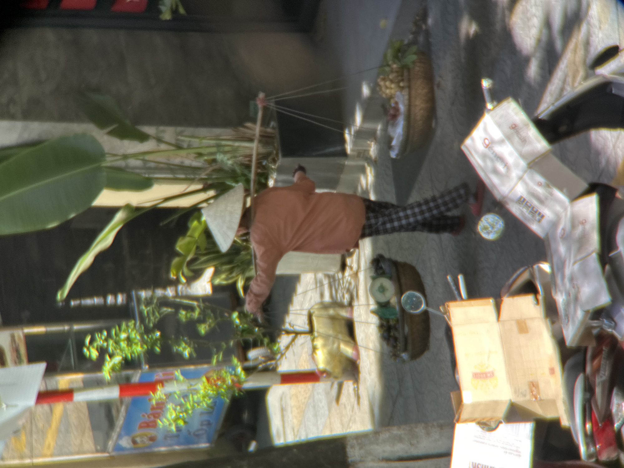 A person wearing a conical hat walks along a sidewalk in Đà Nẵng, Vietnam, carrying a yoke across their shoulders from which two baskets of produce are suspended. The person is wearing an orange shirt and checkered pants. The sidewalk is paved with concrete tiles and there are plants in the background. In the foreground, a motorcycle is parked with several cardboard boxes on it. The lighting suggests it is daytime.