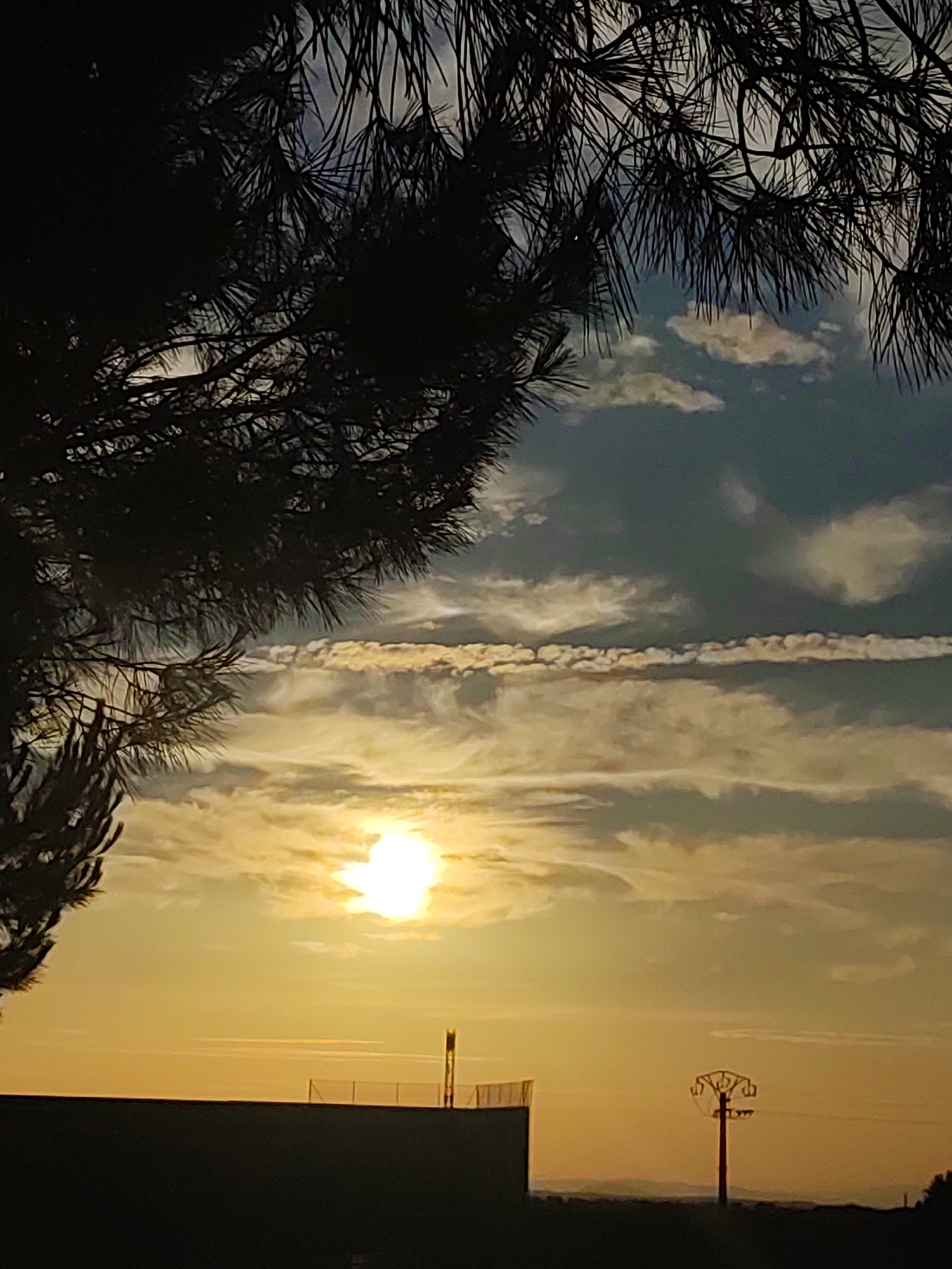 The image depicts a sunset over the city of Sobradillo, Spain. The foreground features the silhouette of pine tree branches against a sky filled with dramatic clouds. The sun, a bright orb, is partially obscured by clouds, casting a warm yellow and orange glow. In the midground, the dark silhouette of a building with a fence is visible, along with a tall, slender utility pole with a distinctive, perhaps decorative, top. Further in the background, the faint outline of rolling hills can be seen against the horizon. The overall scene is serene and picturesque, characteristic of a rural or semi-rural sunset.