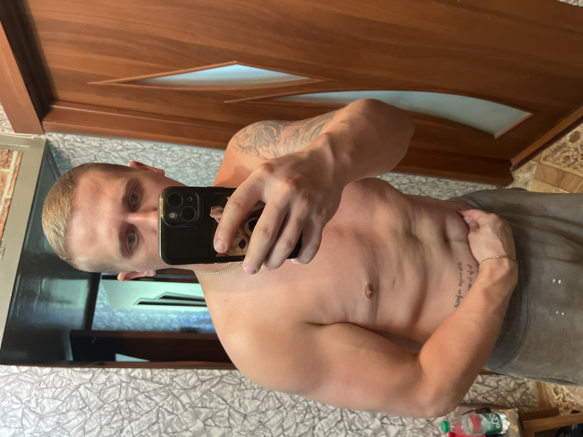 A man with short blonde hair and visible tattoos on his arm is taking a mirror selfie in what appears to be a bathroom. He is shirtless and wearing grey sweatpants. He is holding a black smartphone in his right hand, obscuring part of his face. In the background, a wooden door with a frosted glass insert is visible, as is a textured wallpaper. A bottle of water sits on a small wooden stool to the left of the frame.