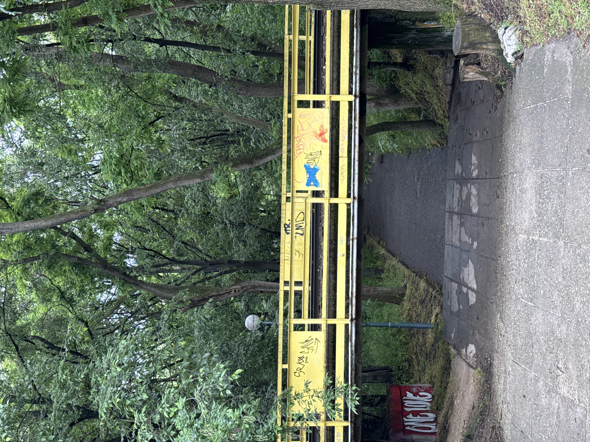 The image shows a yellow metal bridge in Plovdiv, Bulgaria, covered in graffiti. The bridge spans a path and is surrounded by dense trees and foliage. The path is made of concrete. A street lamp is visible near the bridge. A red and white graffiti tag reading "ONE LOVE" is also visible. There are no people in the shot.