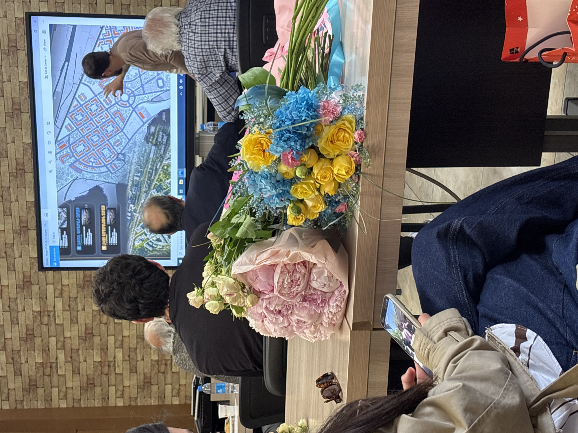The image shows a meeting or presentation in a room with brick walls. Attendees are gathered around a table, and a large screen displays an aerial map and images. A man is pointing at the screen while others watch. A bouquet of blue, yellow, and pink flowers sits on the table. A person is holding a smartphone and looking at the screen. Glasses rest on the table. A red shopping bag is on the table.
Based on the style of the map being discussed on the screen, and the presence of the word "Yerevan" on one of the images, it's likely that the aerial map on the screen is of Yerevan, Armenia. The location within the city cannot be determined from the available information.