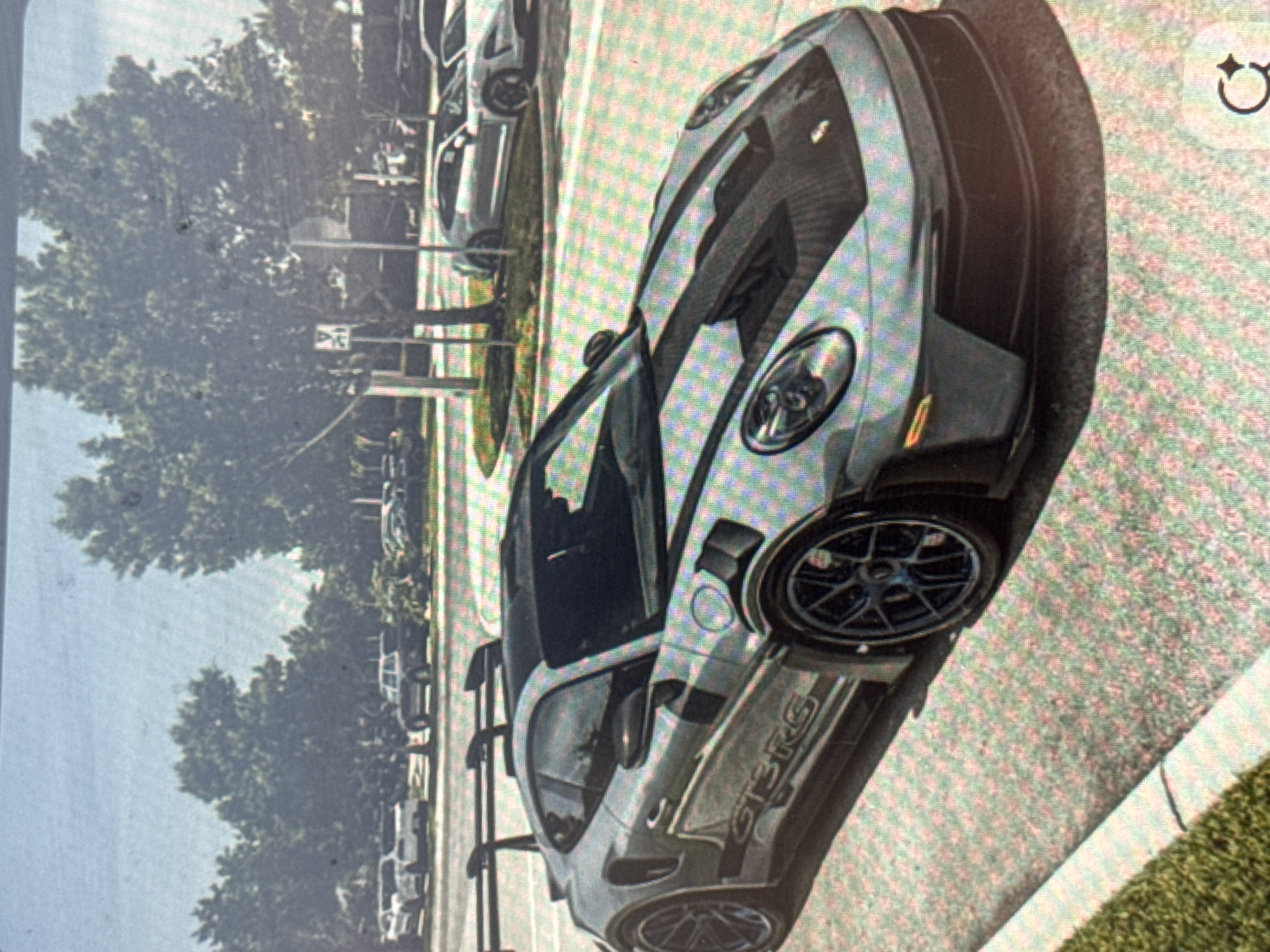 The image displays a grey Porsche 911 GT3 RS sports car parked outdoors during the daytime. The car is angled towards the right, showcasing its front-left quarter, side profile, and rear-left wheel. It features distinctive black accents on the hood, roof, side skirts, and rear spoiler. The car's black multi-spoke wheels are visible, with the front wheel in full view.
In the background, other cars are parked along what appears to be a street or parking lot, lined with numerous green trees. A curb separates the paved area from a grassy verge on the right side of the image. The lighting suggests clear weather conditions.
The primary activity is the car being stationary and parked. No people are visible.
Notable details include the high-performance design of the car, including its large rear wing and aerodynamic elements. The image quality shows a pixelated, grid-like pattern, suggesting it might be a photograph of a digital screen.
Visible text includes "GT3RS" clearly displayed on the lower side panel of the car, identifying its model. There's a small, white rectangular sign in the distant background near the trees, but its text is illegible. While the context of Varna, Bulgaria, is provided, the image itself does not contain any specific visual cues to confirm this location.