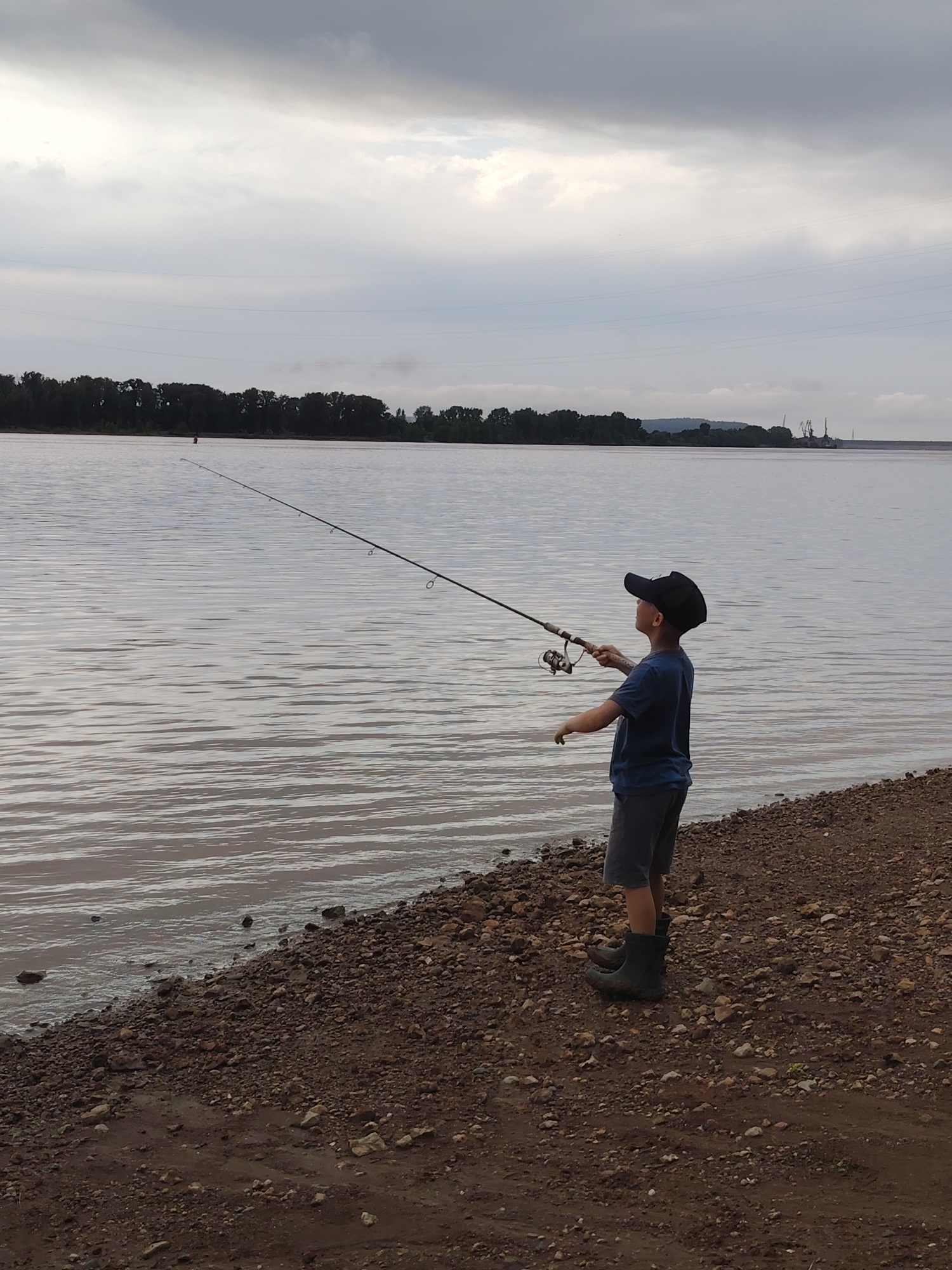 A young boy wearing a dark blue t-shirt, grey shorts, and black rubber boots stands on a gravelly shore, holding a fishing rod and reel. He is facing away from the camera and looking out at a large body of water under an overcast sky. The water is calm with slight ripples. In the distance, a line of trees forms the horizon on the opposite bank of the water. There are industrial cranes visible in the far distance to the right. The scene is suggestive of a fishing outing by the Kama River in the Privolzhsky district of Russia.