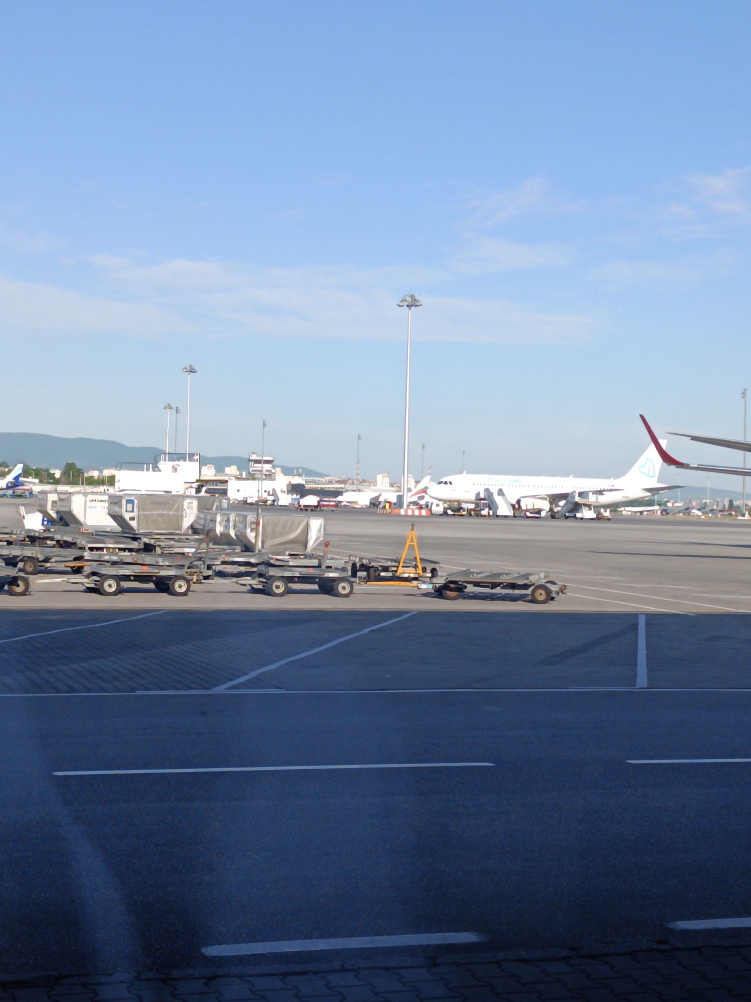The image depicts an airport apron area at Sofia Airport (SOF) in Sofia, Bulgaria. The weather is clear with a blue sky. In the background, there is an Airbus A320-200 with a red vertical stabilizer with an airline logo and a mountain range can be seen in the distance. In the foreground, there are several rows of empty baggage carts parked within painted parking lines on the tarmac. A variety of airport support vehicles and equipment are visible in the mid-ground. Tall lighting poles are evenly spaced in the airport complex. The photograph appears to have been taken from inside the airport terminal, looking out onto the apron.